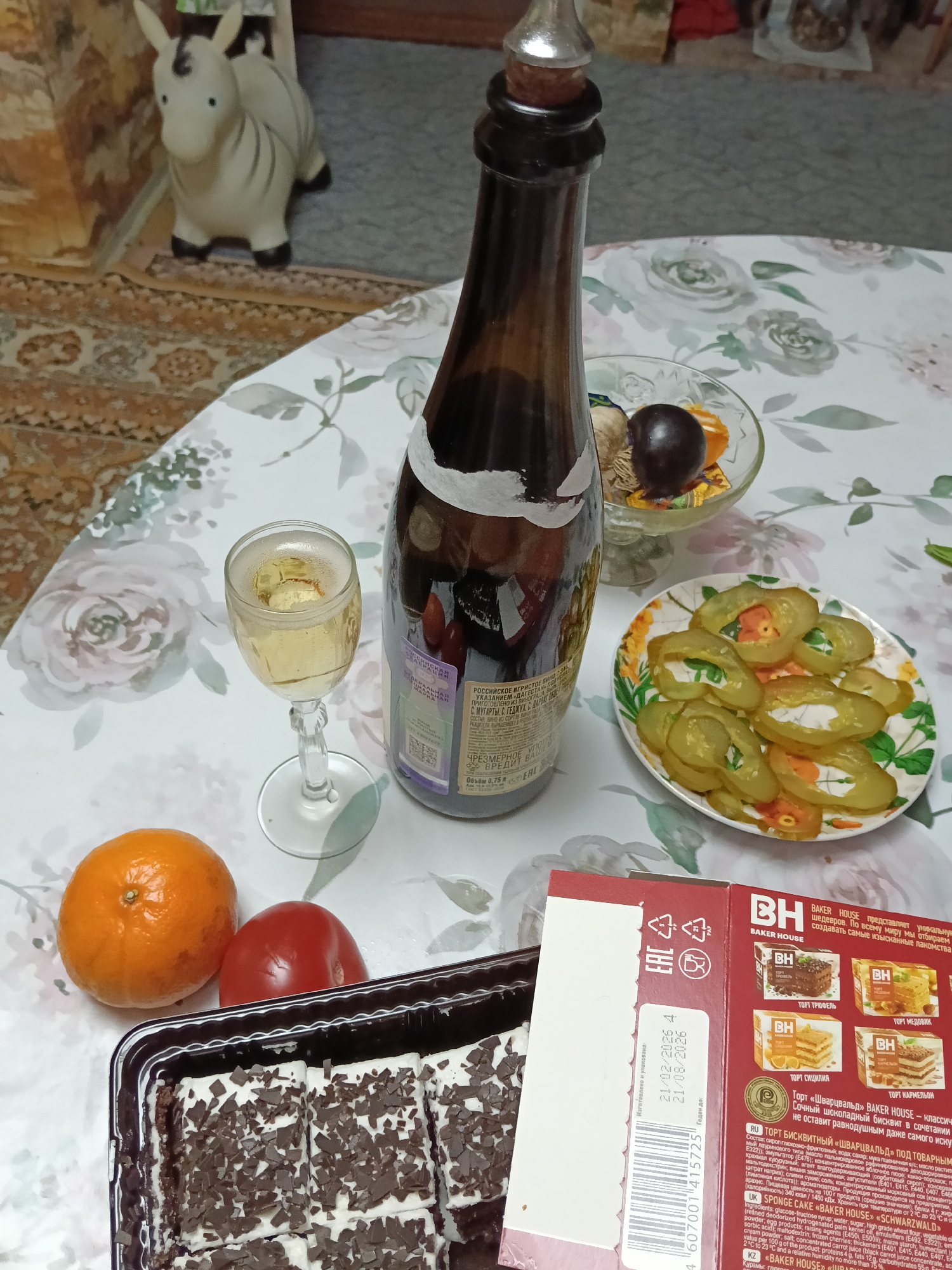 The image shows a table laden with various food items, suggesting a celebratory or casual gathering. The table is covered with a floral-patterned tablecloth. On the table, there is a brown bottle of sparkling wine with a silver stopper, a glass of the same wine, a plate of sliced pickles, a box of "Baker House" sponge cake, an orange, and a tomato. The cake box has a label with nutritional information and expiry dates (21/02/2026 and 21/08/2026). In the background, there is a toy zebra-like animal and indistinct interior details of a room. The location is Красный Маяк, Russia, based on the Cyrillic text on the wine bottle.