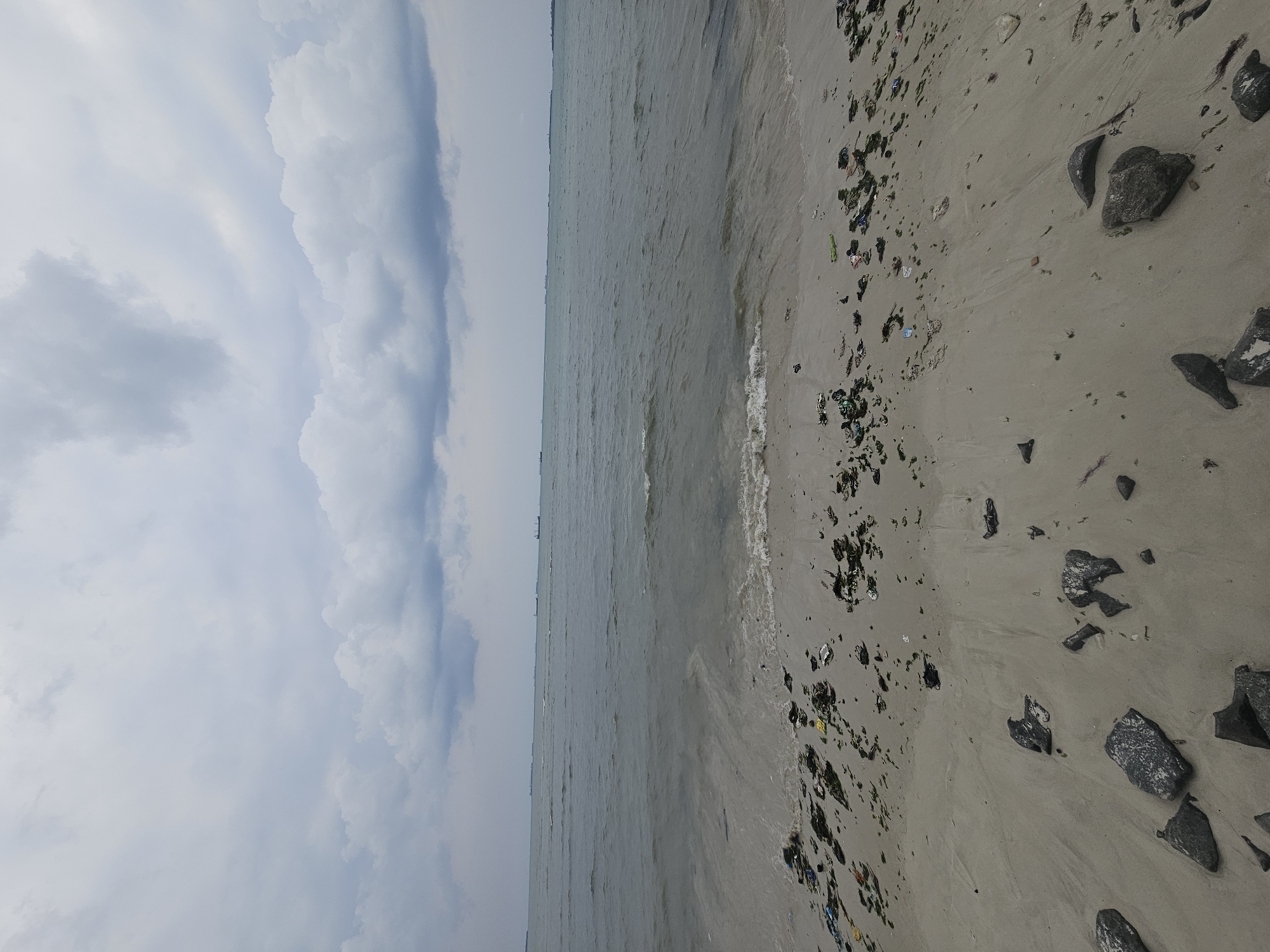 The image depicts a beach scene on a cloudy day. The foreground shows a sandy shore littered with debris, including plastic waste and seaweed, with several dark rocks scattered across the sand. The middle ground features the ocean with gentle waves approaching the shore. In the distance, several large cargo ships are visible on the horizon. The sky is overcast with dramatic, layered clouds. The scene suggests a somewhat neglected coastal area. Based on the context provided, this location is in the city of Dar es Salaam, Tanzania.