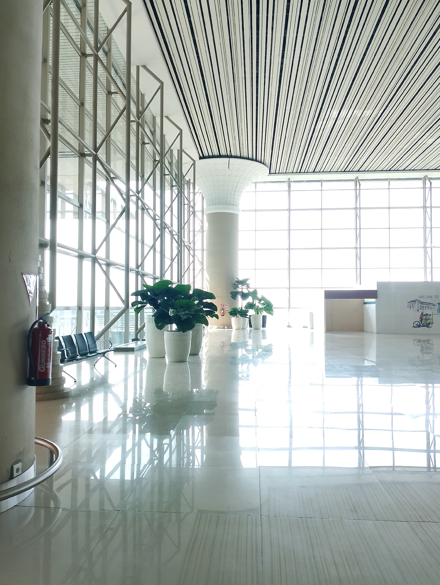 The image depicts an interior view of a modern, spacious building, likely an airport terminal or a large public hall, in Glagah, Indonesia. The architecture features a high, sloped ceiling with white and black linear panels, supported by large, cylindrical pillars and a structural steel frame that forms a grid-like pattern visible through extensive glass windows. Sunlight streams in through these windows, illuminating the polished, reflective floor.
In the foreground, to the left, is a large, beige pillar with a red fire extinguisher mounted on it. Adjacent to the pillar, a row of three black waiting chairs is visible. Further into the scene, several potted green plants in white planters are arranged along the wall, adding a touch of nature to the otherwise minimalist environment.
In the background, a white reception desk or counter is partially visible on the right, with a decorative panel displaying a drawing of a tricycle and the text "WELCOME TO". The overall impression is one of clean lines, bright light, and open space, characteristic of contemporary architectural design. There are no people visible in the image.