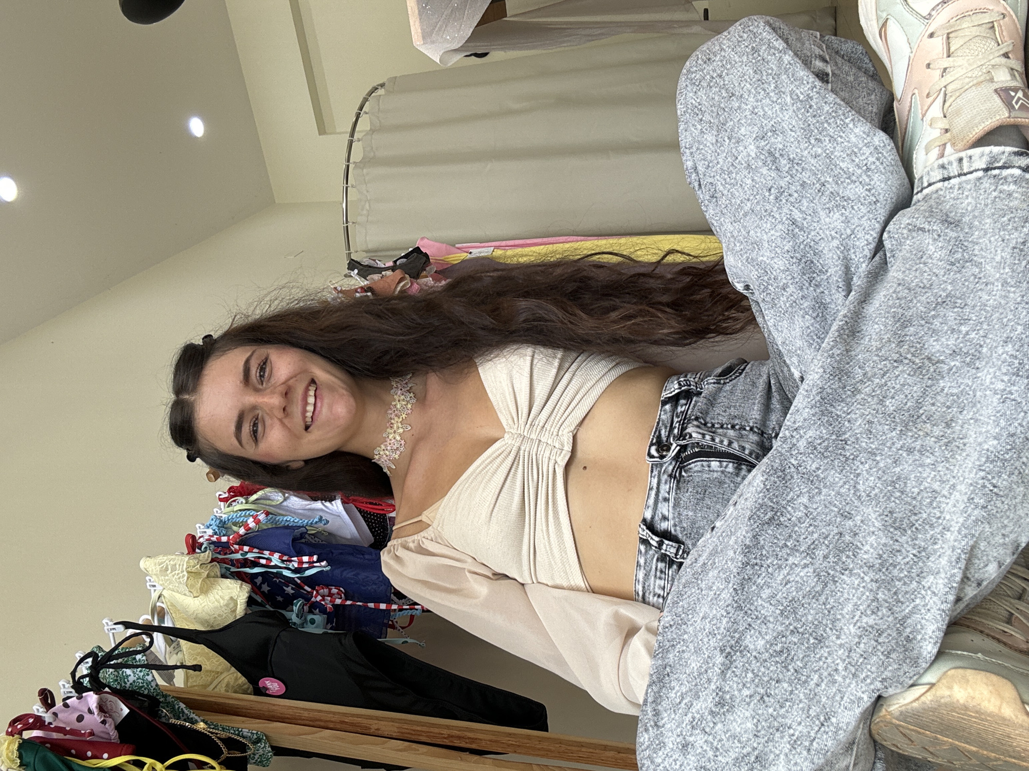 A young Caucasian woman with long, dark, wavy hair and a radiant smile is captured in a full-body, seated shot. She is wearing a cream-colored, ruched, cropped top with sheer, puffed sleeves and a delicate, floral choker necklace. Her lower half is clad in distressed, acid-wash denim jeans. The setting appears to be an indoor boutique or retail space, evidenced by the racks of clothing visible behind her. To her left, a wooden clothing rack displays a variety of colorful swimwear and garments. To her right, a shower curtain-like enclosure hangs, hinting at a changing room. The lighting suggests it is daytime, with overhead spotlights visible on the ceiling. The overall atmosphere is casual and relaxed, with the woman positioned cross-legged on the floor. The presence of a retail environment suggests a location in a commercial area, possibly a street in the city of Nha Trang, Vietnam, which is known for its vibrant shopping scene and fashion boutiques.