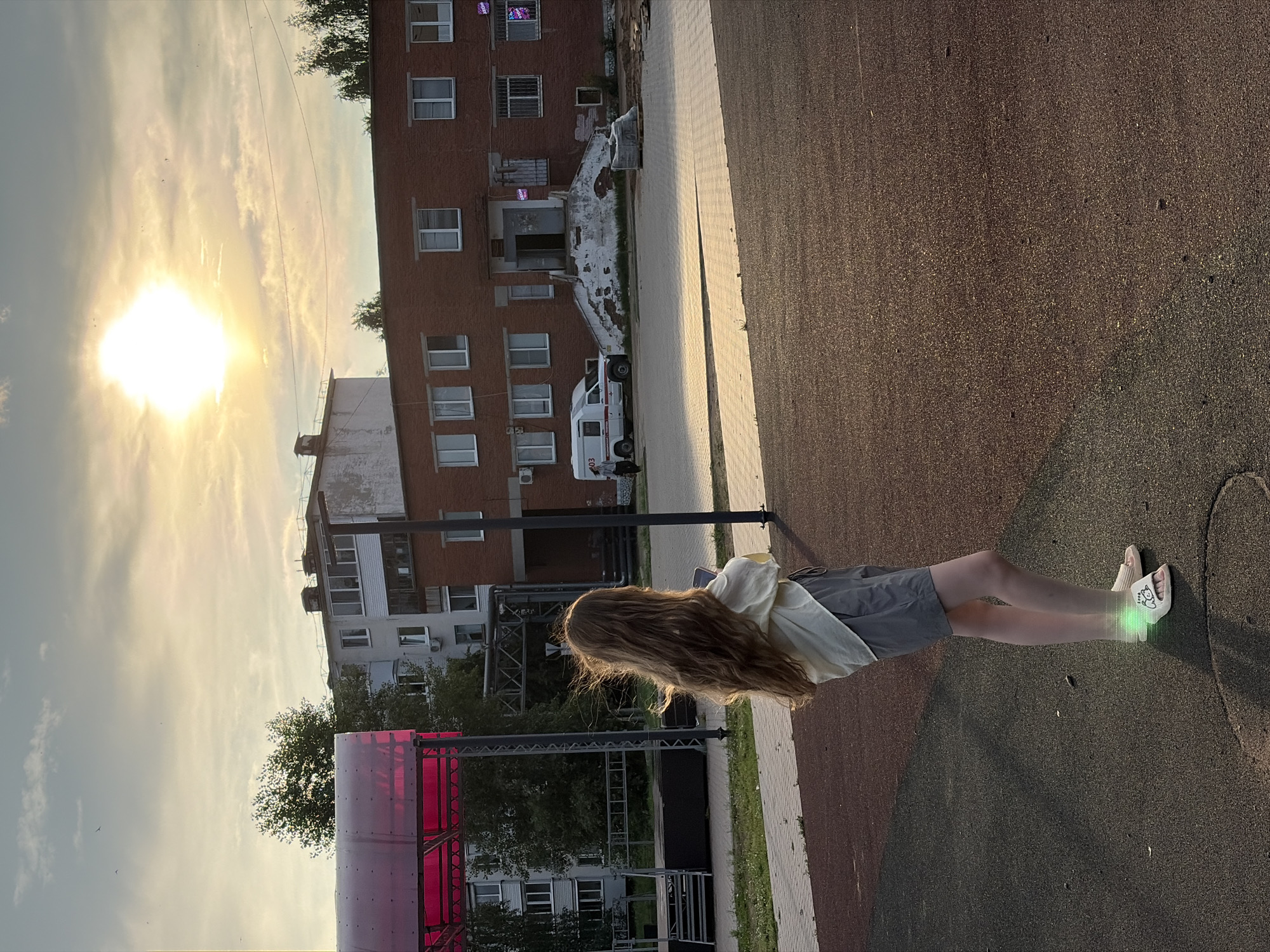 The image shows a girl with long, wavy blonde hair standing in an outdoor area, facing away from the camera. She is wearing a light-colored, loose-fitting top and gray shorts, with sandals on her feet. The ground is a mix of dark asphalt and a reddish-brown textured surface. In the background, there are several buildings, including a brick building with a weathered staircase leading to an entrance, and a multi-story apartment building. A white ambulance is parked in front of the brick building. A pink-roofed structure, possibly a canopy or stage, is visible to the left. The sun is setting, casting a warm glow and creating a hazy sky with some clouds. The environment appears to be a public space within the city of Noviy, Russia.