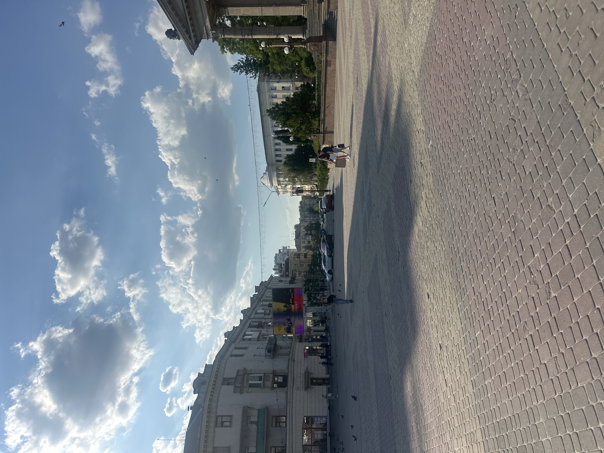 The image shows a public square in Ternopil, Ukraine, on a sunny day with scattered clouds. In the foreground, a paved area with reddish-brown bricks is visible, with shadows cast across it. Several pigeons are scattered on the ground.
To the left, a multi-story building with balconies and shops on the ground floor lines the square. A large digital billboard is mounted on this building. Further down the street, there are more buildings and parked cars.
In the middle ground, a couple walks arm-in-arm across the square. Beyond them, a large, white building with columns and a rounded facade stands, surrounded by trees.
To the right, the corner of another grand building with a portico and columns is visible. Lampposts and trees are also present in this area. The sky is bright blue with fluffy white clouds, and several birds can be seen flying. The overall impression is of a lively urban space on a pleasant day.