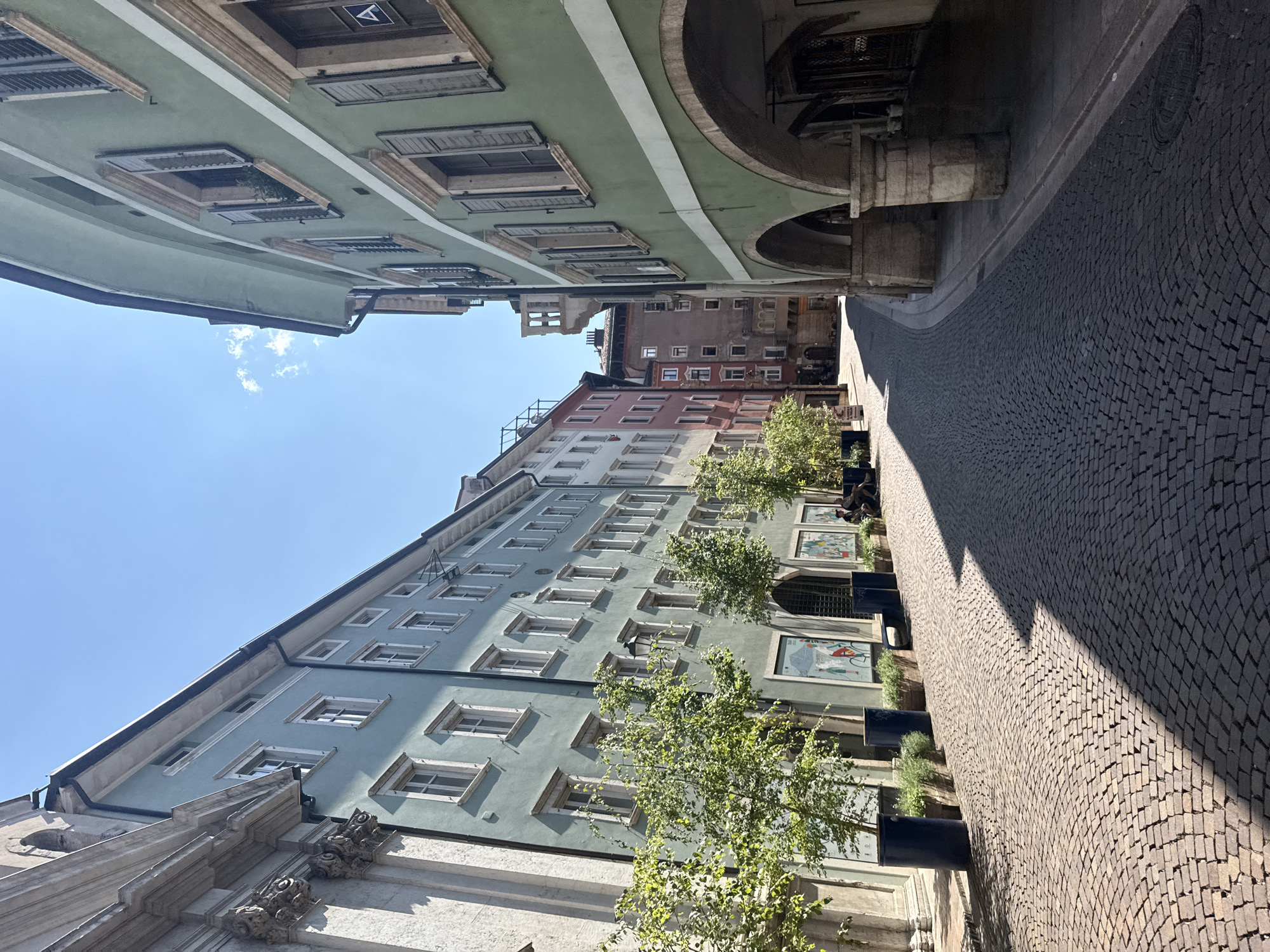 The image captures a sun-drenched cobblestone street in Trento, Italy, winding between buildings with distinct architectural styles. On the left, a building with a pale blue facade features numerous windows with white frames and dark shutters, interspersed with decorative architectural elements. The facade is tall, with multiple stories. Trees in large dark blue planters line the sidewalk, casting shadows on the street. In front of the pale blue building, there are large framed posters.
To the right, a building painted in a muted green hue dominates the foreground. This building has arched ground-floor porticoes supported by stone pillars, offering sheltered walkways. The upper floors have windows with dark shutters and light-colored frames, consistent with the building's color palette. The street itself is paved with uniformly laid dark gray cobblestones, creating a textured surface.
Further down the street, the buildings continue, with a row of pink and red structures visible in the distance, also featuring multiple windows. The sky above is a clear, bright blue with a few wispy white clouds. The lighting suggests it is midday, with strong sunlight creating sharp contrasts between illuminated areas and shadows. Two figures are seated on a bench on the right side of the street, partially obscured by the portico of the green building. The overall impression is of a historic European city street, clean and well-maintained, bathed in sunlight.