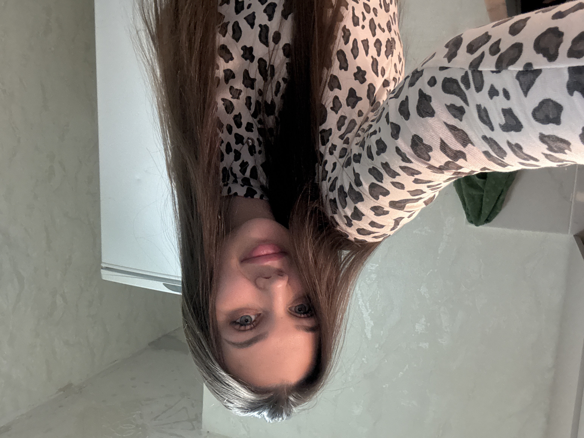 An indoor portrait features a young woman with long, dark brown hair, parted in the middle, looking directly at the camera. She has light-colored eyes, appearing blue or grey, and a subtle, soft smile. Her hair flows around her face and over her shoulders. She is wearing a light-colored top or dress patterned with black and grey animal spots, resembling leopard or cheetah print.
The background consists of plain, light-colored walls, possibly textured or tiled, with a visible corner suggesting an indoor room. A white or light-colored fixture, possibly a shelf or counter, is partially visible behind her head on the right side of the frame. A dark green object, which could be fabric, is also faintly visible in the background on the right. The woman appears to be posing for a photograph, likely a selfie. There is no visible text. The time of day and weather are not discernible from the image.