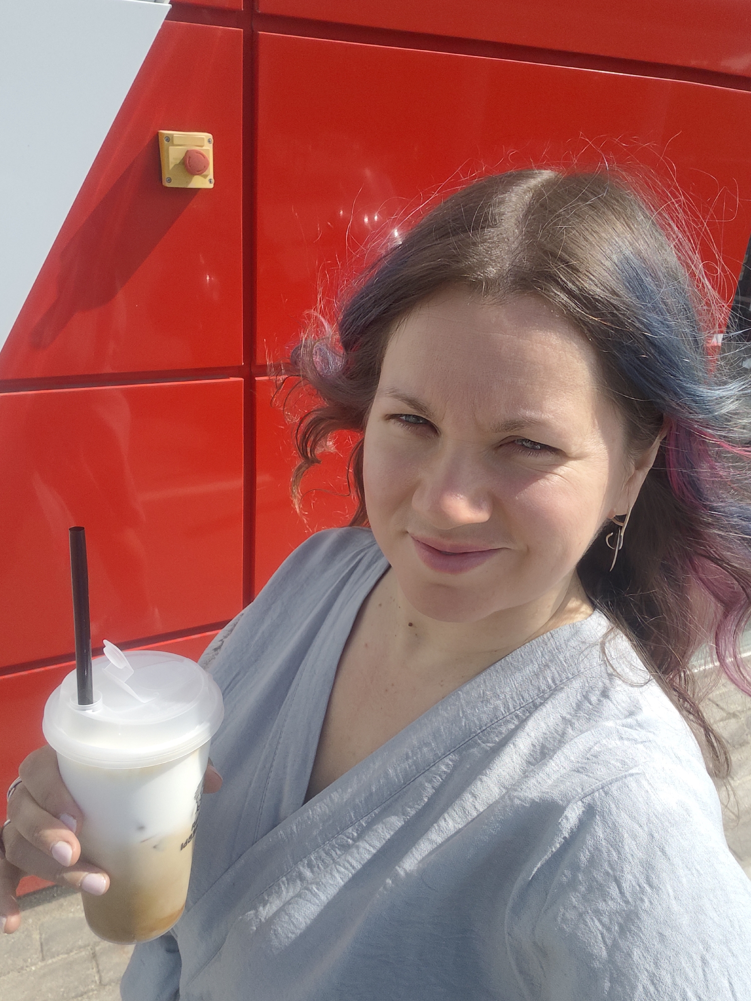 A woman is standing outdoors, holding a plastic cup with a beverage and a straw. She is positioned in front of a red and white building. The woman has brown hair with blue and pink highlights. She is wearing a light blue top. A yellow box with a red button is on the red building. The image was taken in Sholokhovo, Russia.