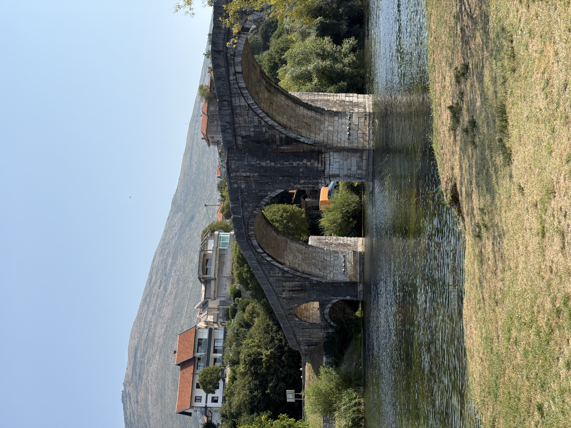 The image captures a historic stone bridge spanning a calm river in Trebinje, Bosnia and Herzegovina. The bridge, a prominent architectural feature, is constructed with large, weathered stone blocks and features multiple arches, with the largest central arch dominating the composition. The river's surface is rippled, reflecting the clear blue sky and the bridge's stone structure.
On the riverbank in the foreground, dry, sun-bleached grass covers the ground, with patches of greener growth scattered throughout. Behind the bridge, on the elevated embankment, are several modern houses with red-tiled roofs, interspersed with lush green trees and foliage. The houses appear to be residential properties, some with balconies and large windows.
In the background, a large, sparsely vegetated mountain slopes upwards, its contours softened by distance. The sky is a clear, unbroken expanse of light blue, indicating a bright, sunny day. The overall scene evokes a sense of tranquility and historical presence, blending ancient architecture with contemporary life within a natural landscape. There are no people visible in the image.