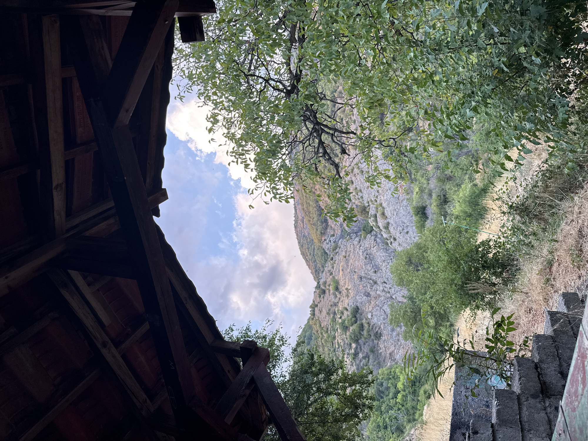 The image presents a view from beneath a rustic wooden structure, likely a gazebo or shelter, looking out towards a mountainous landscape. The foreground is dominated by the dark, weathered beams and tiled roof of the structure. Through the opening, a dry, rocky mountainside covered in sparse vegetation and trees is visible, under a cloudy sky. To the right, a leafy green tree branches into the frame, partially obscuring the view. In the lower left, a series of concrete steps or blocks, showing signs of age and wear, are partially visible. The overall impression is one of a natural, somewhat overgrown setting, possibly a resting spot in a rural or mountainous area. The location is identified as Sopot, Bulgaria.