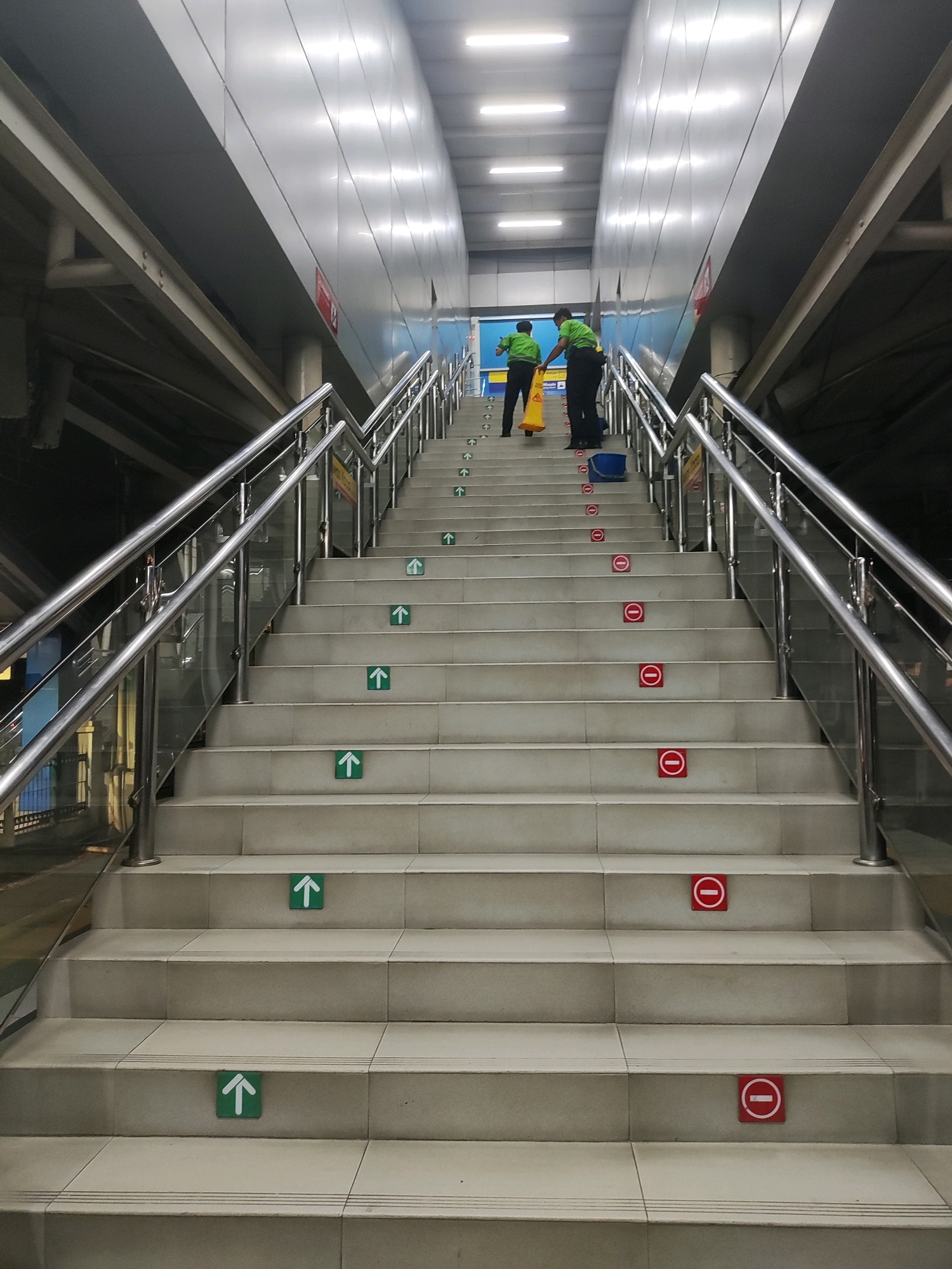 In East Jakarta, Indonesia, two cleaners in green shirts and dark pants are on a wide, outdoor staircase. One cleaner holds a yellow cone, and the other has a blue bucket. The staircase is lined with stainless steel railings and glass panels. The steps are marked with green arrow stickers pointing upwards and red circular stickers with a horizontal line, indicating "do not enter" or "no entry." The cleaners appear to be working, potentially cleaning or marking the stairs. The environment seems to be a public space, possibly a transit station or an elevated walkway.