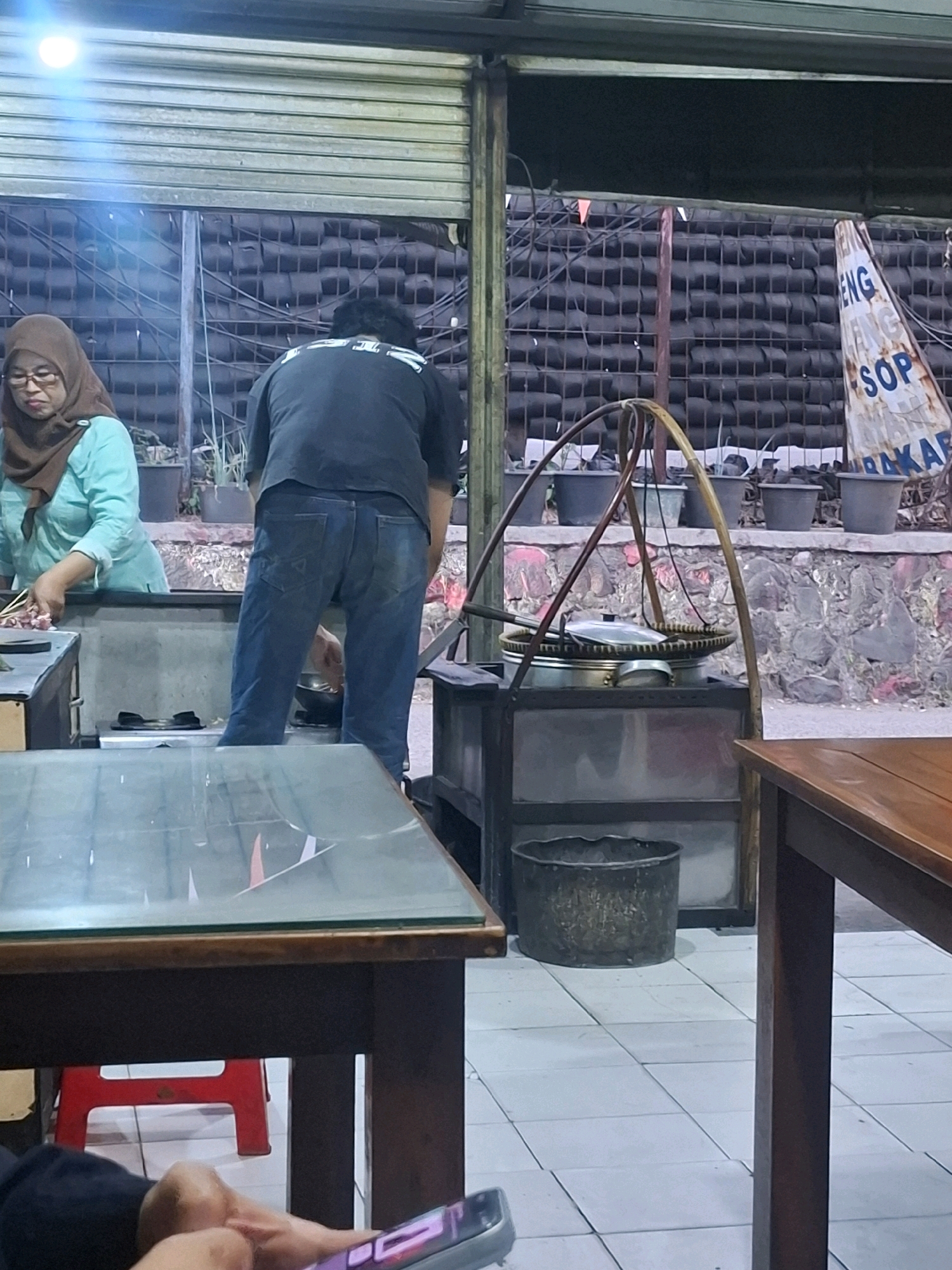 An outdoor food stall in Citeureup, Indonesia, features two individuals preparing food. A woman wearing a brown hijab and glasses stands to the left, tending to food on a stove. A man in a black t-shirt and blue jeans is positioned behind her, also engaged in cooking. The stall is equipped with a portable stove, pots, and cooking utensils. In the background, a wall is adorned with a sign that reads "ENG SOP BAKAR." Tables and chairs suggest a dining area. The environment appears to be a street-side establishment, with an overhead rolling door in the background.