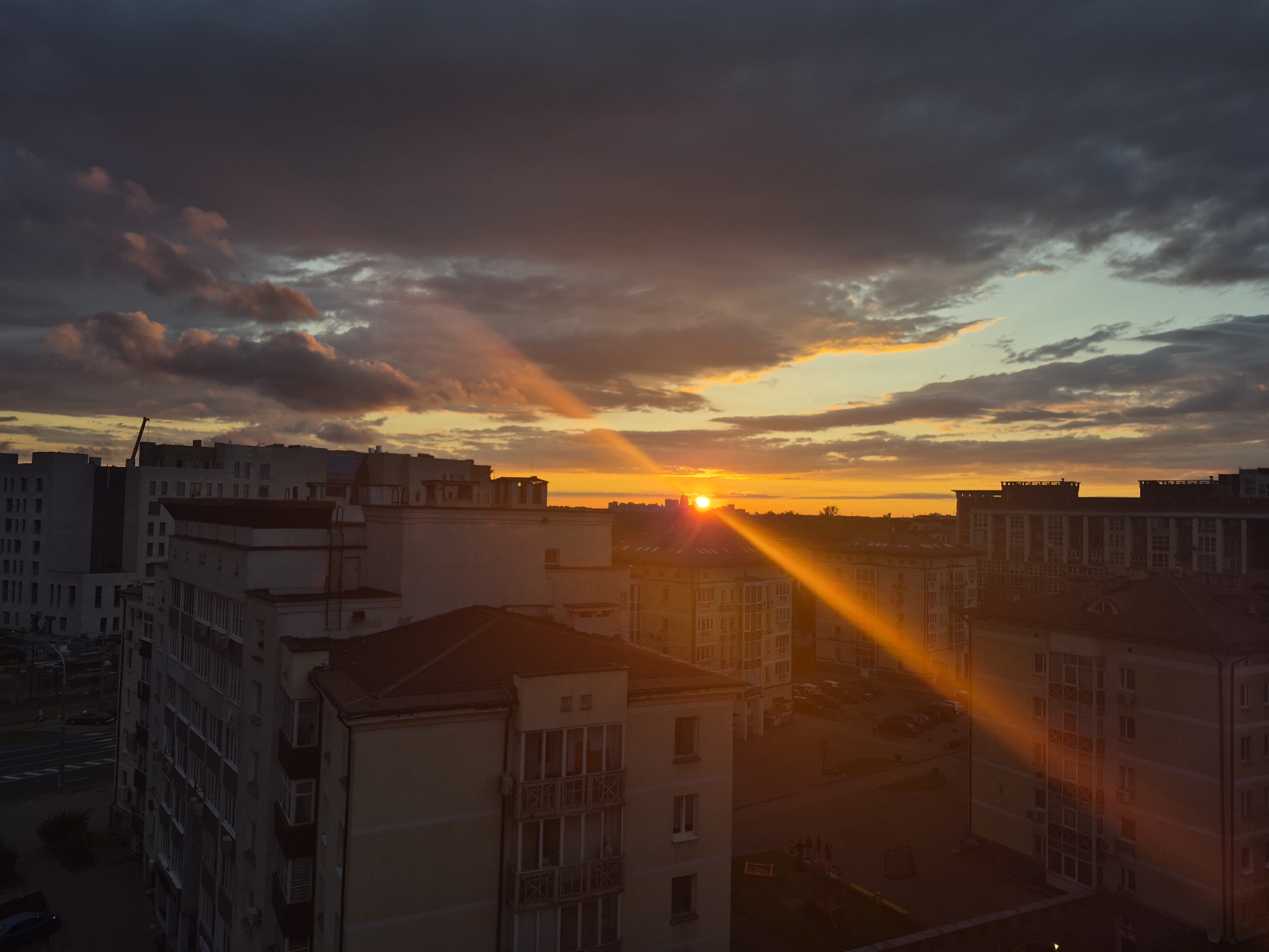 An aerial view captures the dramatic interplay of light and shadow over a cityscape during sunset. The sky is a canvas of dark, heavy clouds tinged with hues of orange, yellow, and a faint teal where the sun's rays pierce through. The setting sun, a bright orb on the horizon, casts long, golden beams that cut through the atmosphere, illuminating parts of the urban landscape.
In the foreground, a cluster of residential buildings, likely apartment blocks, dominate the scene. Their facades are a mix of cream, beige, and grey, with numerous windows reflecting the fading light. Some buildings feature balconies and exposed architectural details. The architecture suggests a post-Soviet era construction.
Below the buildings, streets and parking areas are visible, with a few cars parked. A playground with colorful equipment is discernible in one of the courtyards. The overall impression is that of a residential district in a city.
While the specific location is not explicitly indicated in the image itself, the architectural style of the buildings and the general urban layout are consistent with areas found in Minsk, Belarus. The scene depicts a quiet, possibly evening, moment in a residential part of the city, characterized by the striking natural phenomenon of a sunset. No people are clearly visible in the image.