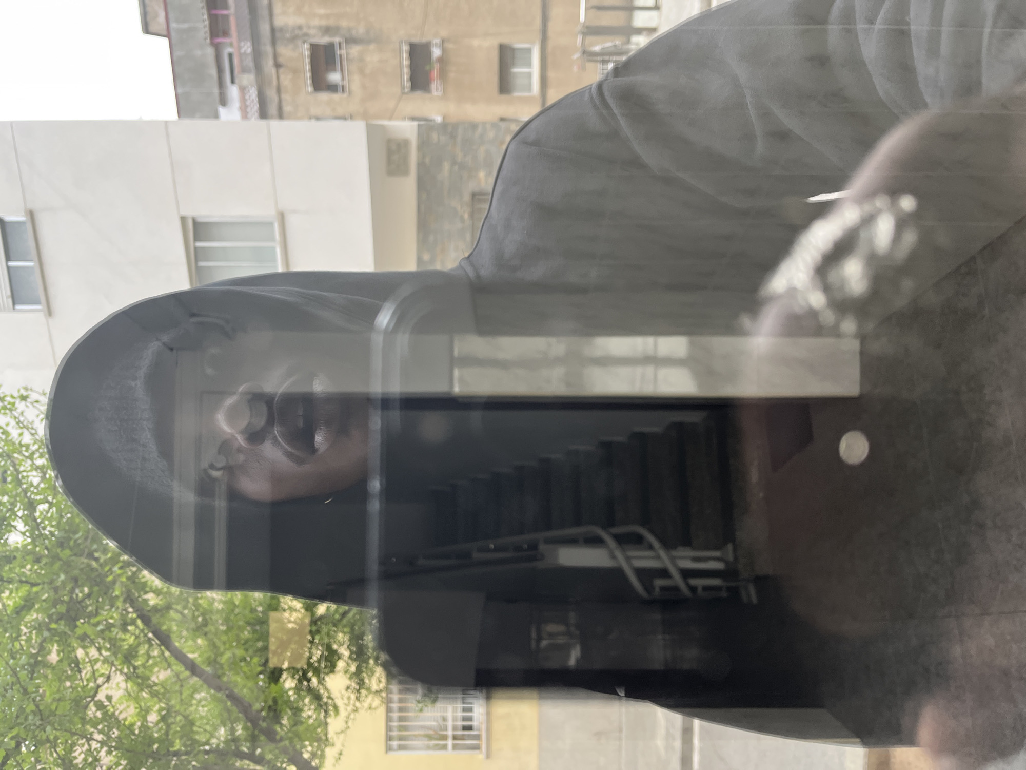 A reflection in a glass window reveals a man in a hooded sweatshirt standing indoors. The reflection superimposed on the man shows an exterior scene, including a stairway, trees, and surrounding buildings. The urban architecture suggests a location in Barcelona, Spain.