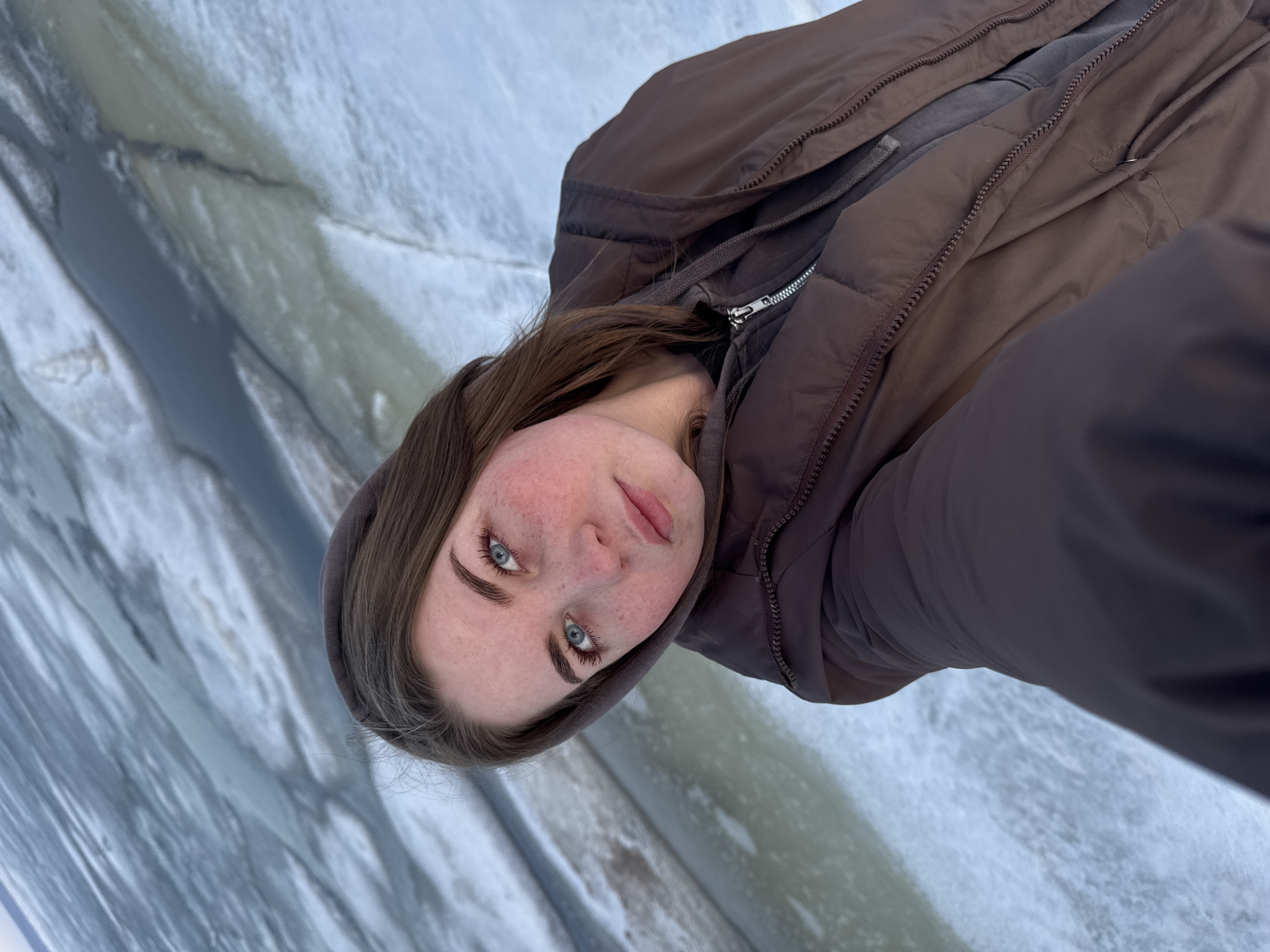 A young woman with fair skin, freckles, and blue eyes is the central subject of this photograph. She is wearing a brown hooded jacket and a gray sweatshirt underneath. The jacket is zipped up. Her hair is brown and partially covers the hood. The background appears to be a frozen body of water with visible cracks and patterns on the ice.
While the user requested the location of "Noviy, Russia," the image does not provide enough forensic detail to determine the specific geographic location. It only shows a person and a frozen body of water, which are not unique features of any specific place.
