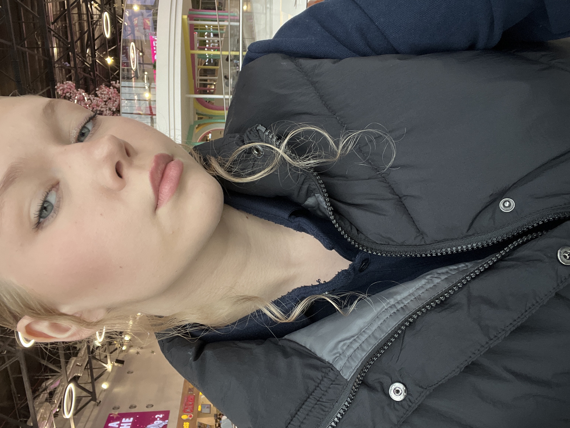 The image is a close-up, slightly angled, outdoor selfie of a young Caucasian woman with blonde hair and blue eyes. She is wearing a dark blue collared shirt and a black puffer vest. Her hair is partially styled in curls that fall around her face and neck. The background appears to be an indoor public space, possibly a mall or entertainment center, with modern architecture, including suspended lighting fixtures, railings, and colorful structures in the distance. One of the background elements suggests it is the city of Perm, Russia, specifically the entertainment zone of the "Planeta" shopping and entertainment complex. The woman's expression is neutral.
