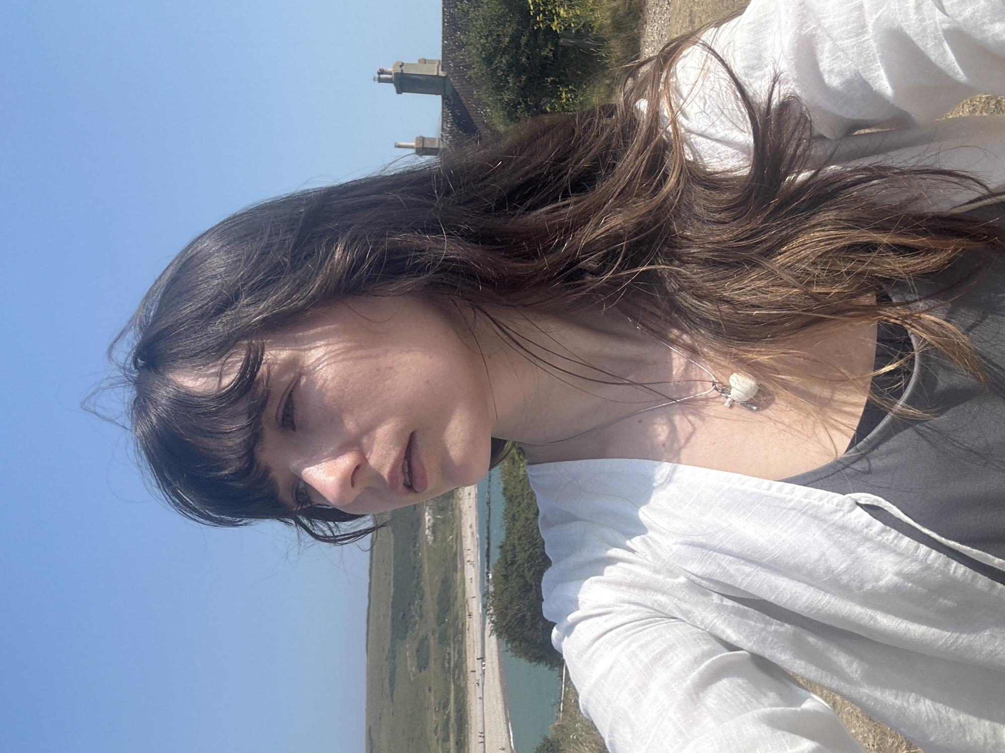 A young woman with long, dark, wavy hair and bangs is shown in a close-up, outdoor selfie. She is looking to her right, with her head tilted slightly upwards. She is wearing a white, open-fronted shirt over a dark gray tank top. A silver necklace with a white, heart-shaped pendant hangs around her neck.
The background reveals a coastal landscape under a clear blue sky. A pebbled beach meets a body of calm, turquoise water, with several small figures visible on the beach. Rolling green hills rise behind the beach. In the middle ground, a red-tiled roof with two prominent brick chimneys is visible, suggesting a building situated on the hillside. The environment appears sunny and warm, typical of a seaside location. The image was taken in Seaford, United Kingdom.