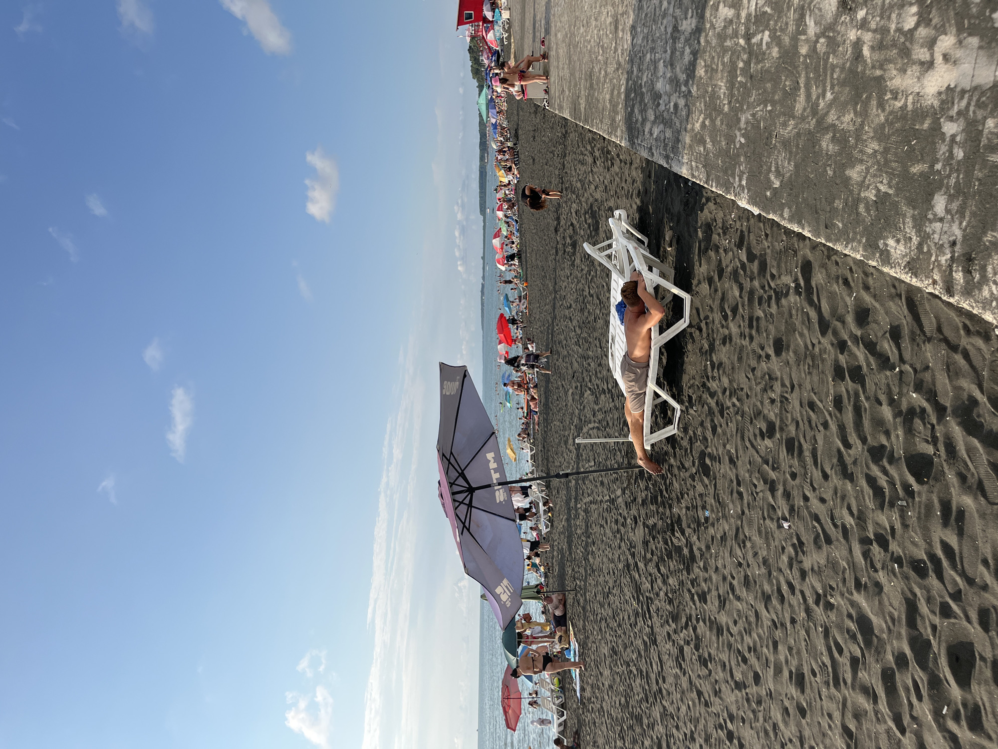 The image depicts a crowded beach scene in Ureki, Georgia, characterized by dark volcanic sand and a calm sea. In the foreground, a man lies on a white lounge chair, facing downwards, wearing light-colored shorts and a blue cap. To his left, another lounge chair is empty. Further into the scene, numerous people are scattered across the beach, many reclining on lounge chairs or standing under colorful beach umbrellas. The sea is visible in the background, with a clear blue sky above featuring scattered white clouds. The concrete promenade runs along the right side of the beach, separating it from the sand. The overall atmosphere suggests a sunny day at a popular tourist destination.
