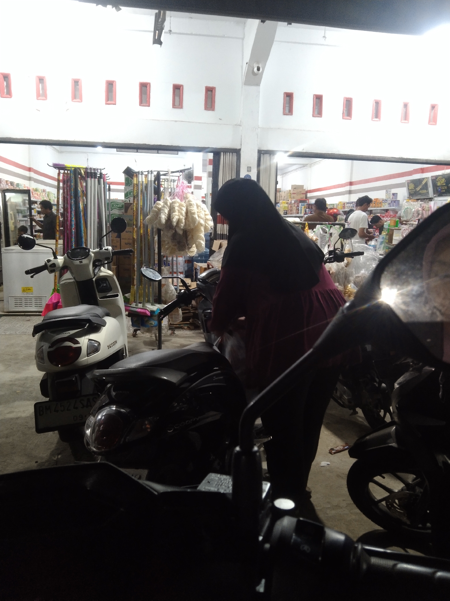 This is a medium shot, taken at night, of a street-level shop in Langkai, Indonesia. The shop appears to sell general goods and groceries, with shelves stocked with various items visible in the background. Several motorcycles are parked in front of the shop, one of which is predominantly in the foreground, obscuring some of the shop's facade.

In the foreground, a woman wearing a black headscarf and a dark pink dress is standing near a white scooter. She appears to be interacting with the scooter or items placed on it. To the right of the woman and scooter, another dark-colored motorcycle is partially visible.

In the background, several people are seen inside the shop. One man is near a refrigerator on the left, and other individuals are browsing the shelves. The shop is illuminated by artificial lighting, creating a well-lit interior. The exterior of the building features a white facade with several small, rectangular windows with red frames. The ground is concrete.