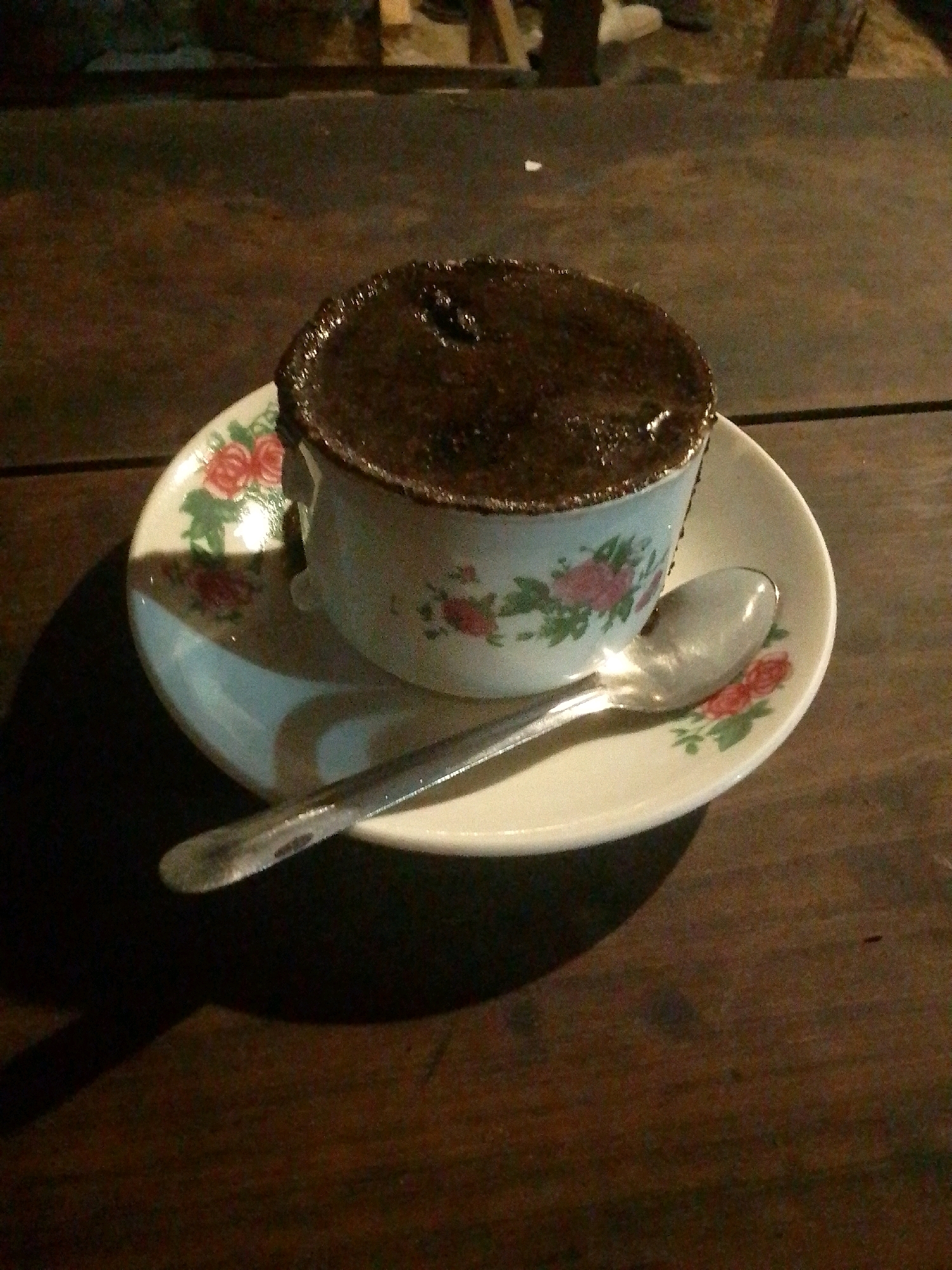 A close-up shot shows a cup of coffee on a saucer, with a spoon placed on the saucer. The cup and saucer are white with a floral pattern of red roses and green leaves. The coffee appears dark and frothy. The scene is set on a wooden table, and a shadow from the cup and saucer is cast to the left. The background is dark and out of focus, suggesting an indoor or dimly lit outdoor setting. The image appears to be taken in Trenggalek, Indonesia, though there are no specific visual cues to confirm this location within the frame itself.