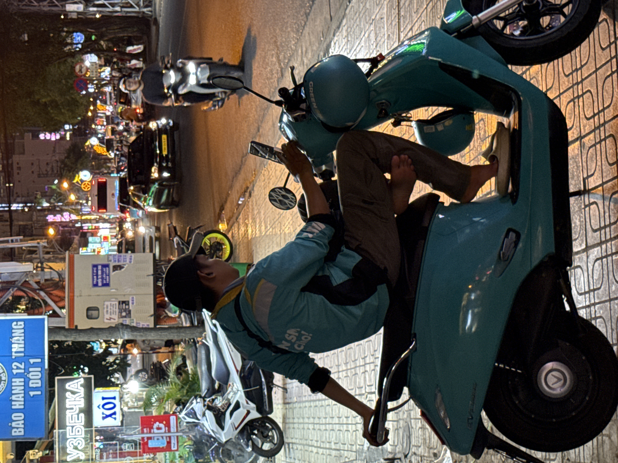 At night on a busy street in Nha Trang, Vietnam, a person is sitting on a teal scooter. The person is wearing a teal jacket with yellow accents, brown pants, and a black baseball cap. Their left arm is resting on the handlebar of the scooter, and they are holding a smartphone in their right hand. A teal helmet is hanging from the side of the scooter. The street is lined with buildings, traffic lights, and other vehicles. The scooter has a "GREEN" logo on the side. In the background, other motorbikes and cars are visible, as well as streetlights and illuminated signs. The ground is paved with interlocking pavers. The image captures a moment of urban life at night.