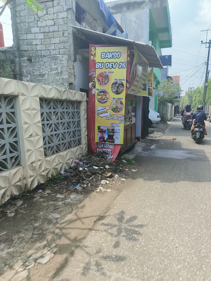 The image shows a street scene in Padalarang, Indonesia (latitude: -6.858, longitude: 107.483). A large, colorful sign advertising "Bakso Bu Dev 26" is displayed in front of a brick building. The sign lists various menu items including "Mie Bakso", "Mie Kocuk", "Mie Ayam", and "Mie Jamur". The sign also advertises a "Promo Paket" for 13,000 rupiah. A pile of debris, likely garbage, is located in the foreground near the building. A motorcycle is visible parked on the right side of the street, and there is a small, light-colored car partially visible behind the sign. Shadows from trees are cast on the pavement, and the overall environment appears to be a residential or commercial area with a mix of businesses and homes. There are no people visible in the image.