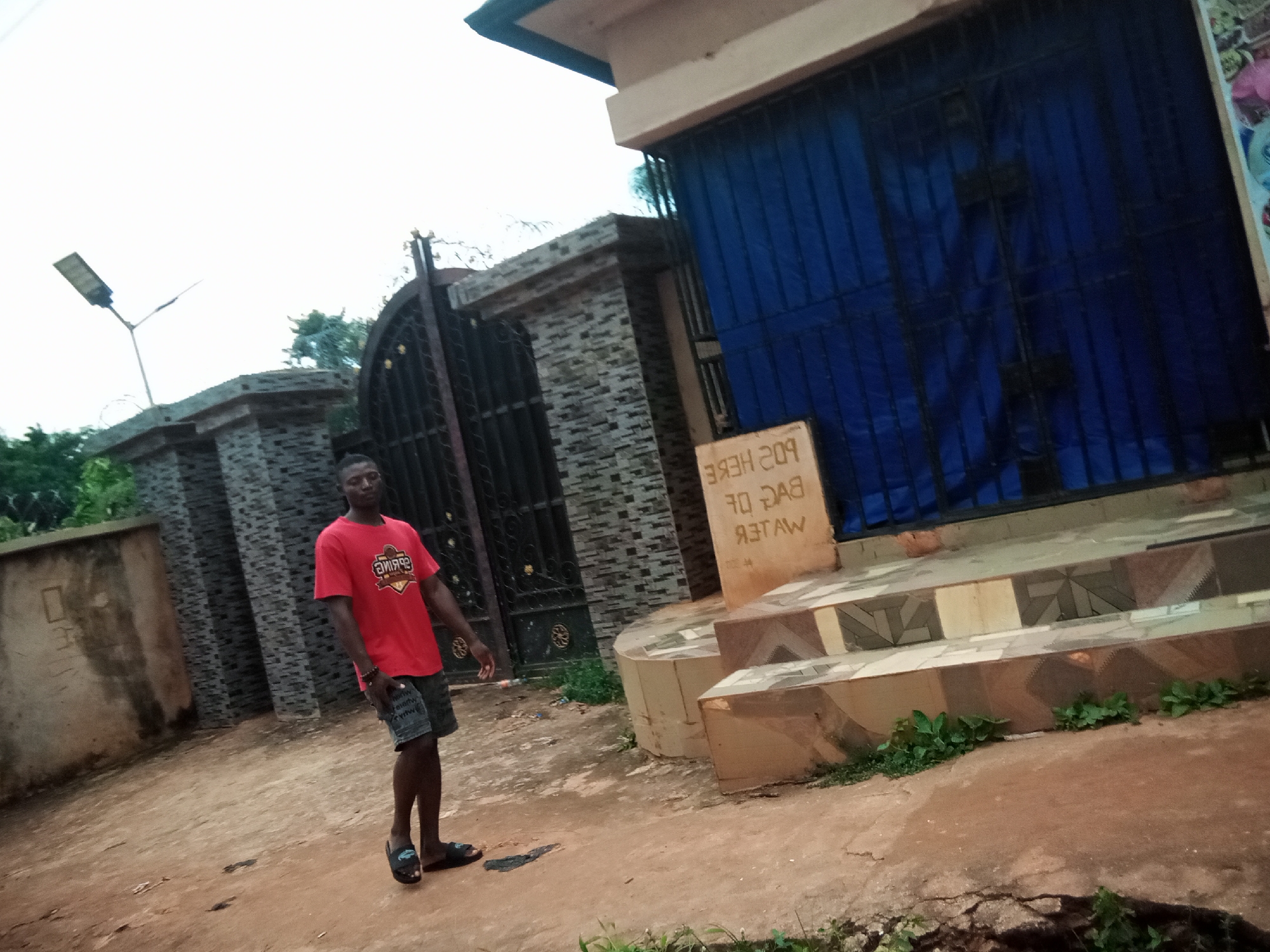 In Oko Town, Nigeria, a young man wearing a red t-shirt and denim shorts stands on a dirt path in front of a gate. The gate is made of black metal bars and is partially obscured by a blue tarp. To the left of the gate is a stone wall. A sign is posted on the wall, facing the viewer. The sign has text written in white. In the background, a street lamp is visible. The ground is a mix of dirt and some patches of greenery. The sky is overcast.