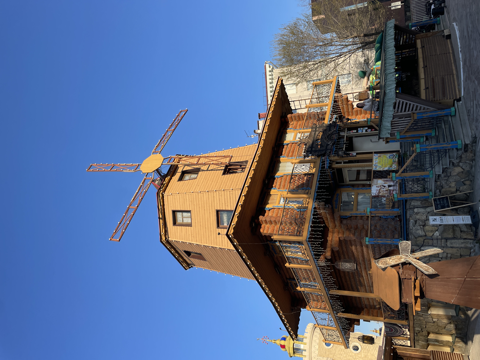 A low-angle, wide-shot captures the exterior of a building shaped like a windmill, located in Kazan, Russia. The primary structure is constructed of brown wood. The building features a non-functional windmill with four orange blades extending from the top. The windows are visible along the sides of the windmill structure. The building has multiple stories, with balconies decorated with ornate metalwork and small light bulbs. The lower portion of the building includes a mix of wood and stone construction, adorned with various decorative elements and signage. The background consists of parts of other buildings, a tree without leaves, and a clear blue sky.