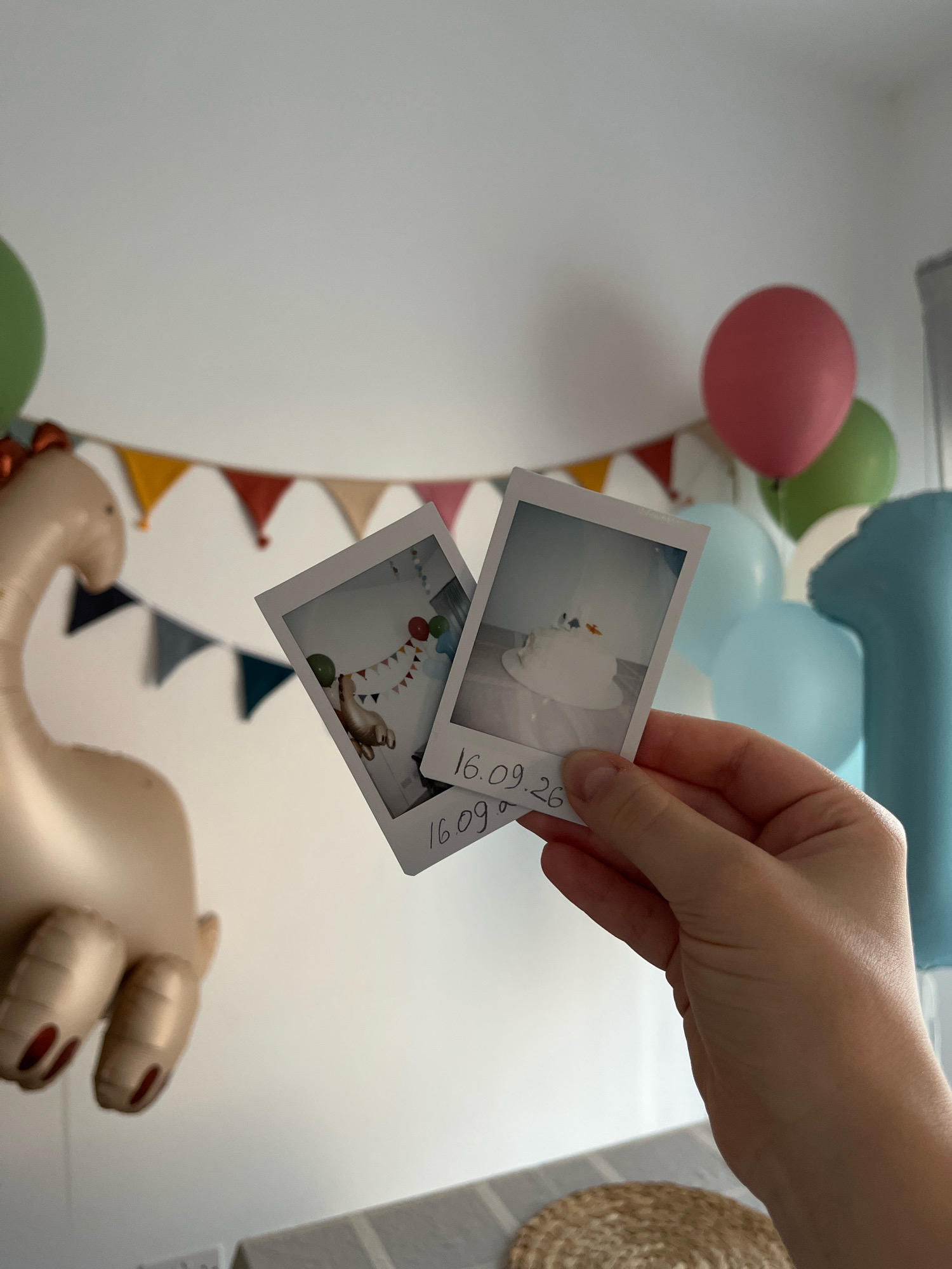 The image captures a close-up of a hand holding two instant photographs against a blurred background of a room decorated for a celebration. The photographs appear to be of a birthday cake and a balloon animal, with dates written on them: "16.09.26" and "16.09". The background features several balloons of various colors, including pink, green, and blue, as well as a festive banner composed of triangular flags. A large, golden dinosaur-shaped balloon is partially visible on the left side of the frame. The scene suggests a birthday party setting, possibly for a child, given the presence of the cake, balloons, and a dinosaur balloon. The location is specified as Limassol, Cyprus, though no specific landmarks or contextual clues within the image directly confirm this location. The lighting appears to be natural, coming from an unseen source, casting soft shadows. The hand holding the photographs has neatly painted fingernails.