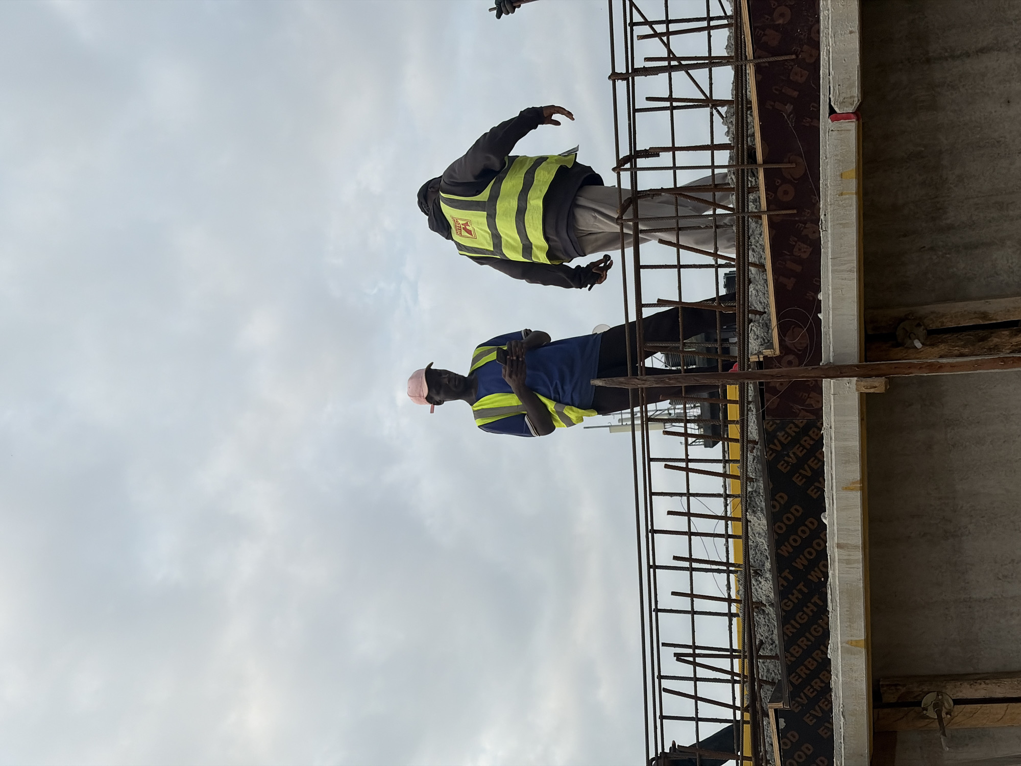 Two construction workers are visible on a structure under construction against a cloudy sky. The worker on the left, wearing a pink cap and a yellow safety vest over a blue shirt, is looking down at a mobile phone. The worker on the right, facing away, is wearing a dark hooded jacket with a bright yellow safety vest over it. They are standing on a platform reinforced with rebar and plywood. The lower portion of the image shows concrete structures and wooden supports. The scene is likely a construction site in Alaguntan, Nigeria, indicated by the context.