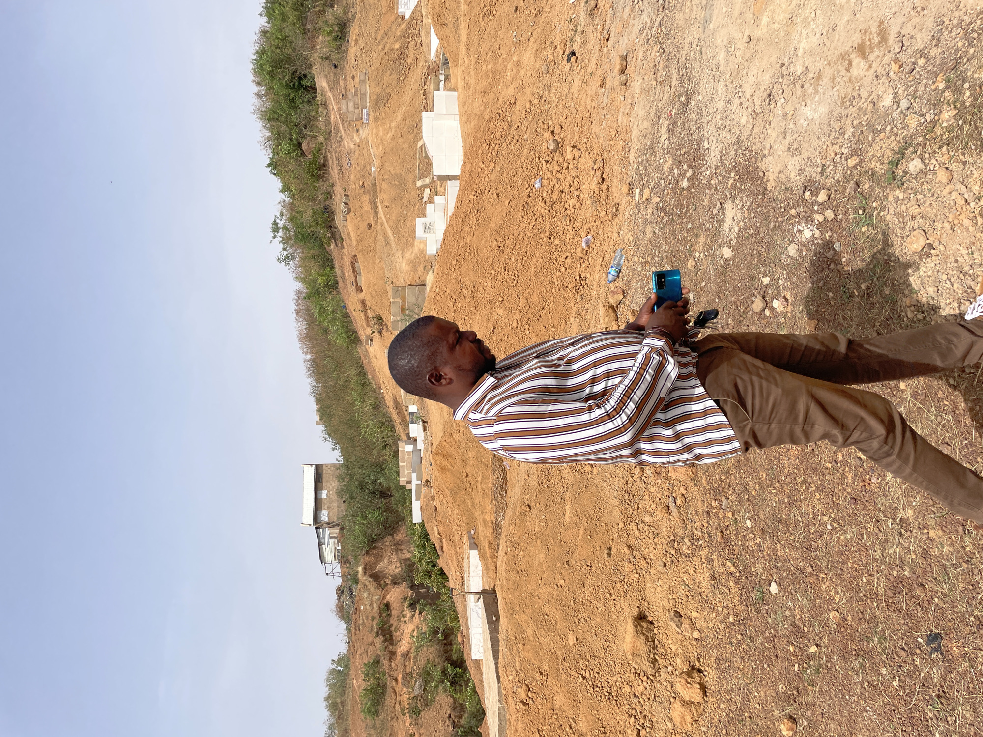 A man stands on a dirt slope in Bar Jirgi Summa, Nigeria. He is using a blue mobile phone. The slope is part of a hill that features vegetation and concrete structures, possibly graves or building foundations. The sky is pale. The man wears a horizontally striped shirt, brown pants, and white shoes. The ground is uneven with dirt and small stones.