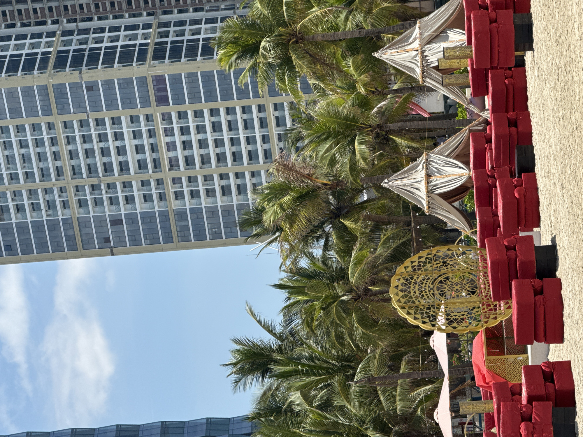 The image shows an outdoor setting in Đà Nẵng, Vietnam, featuring a beachside event. Tall palm trees sway in the foreground, interspersed with decorative elements like a large gold, filigree-style hanging ornament and several small, tent-like canopies with beige fabric. Rows of stacked red cushions line the right side, likely for seating. The background is dominated by a large, modern building with a grid pattern of windows, typical of urban architecture. The sky is blue with a few scattered clouds. There are no people visible.