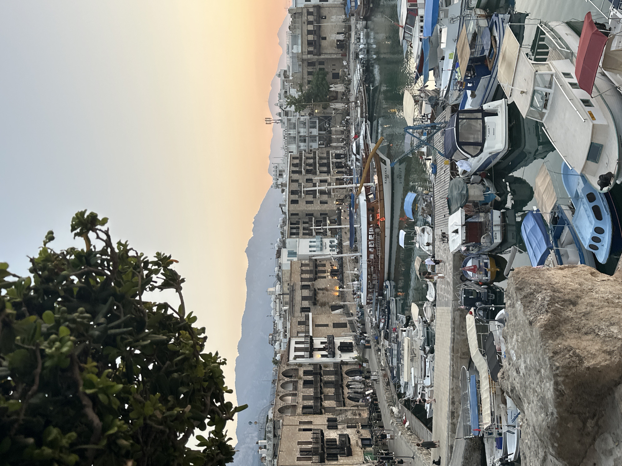 The image depicts a panoramic view of Kyrenia's harbor in Northern Cyprus, captured during the golden hour. The foreground is dominated by a large, rough-hewn rock formation in the bottom left corner. Beyond the rock, a multitude of boats of various sizes and types are moored in the calm harbor waters, reflecting the warm, soft light of the setting sun. Many of these boats are small motorboats, while a larger, traditional wooden sailing vessel with multiple decks and sails is prominently positioned near the center of the harbor.
The middle ground showcases the picturesque Kyrenia harbor, lined with historic stone buildings. These buildings feature arched windows, balconies, and dark wooden shutters, conveying a sense of age and architectural character. Restaurants and cafes with outdoor seating are visible along the waterfront, with a few patrons seated at tables. People are seen walking along the harbor promenade.
In the background, a range of modern white buildings and a few taller structures with antennas rise behind the older architecture, indicating the city's development. The distant mountains form a soft, hazy silhouette against the pale orange and yellow hues of the sunset-lit sky. A single green tree branch with dense foliage extends into the top left corner of the frame, partially obscuring the sky and adding a natural element to the urban and maritime scene. The overall atmosphere is serene and tranquil, characteristic of a peaceful evening in a coastal city.
