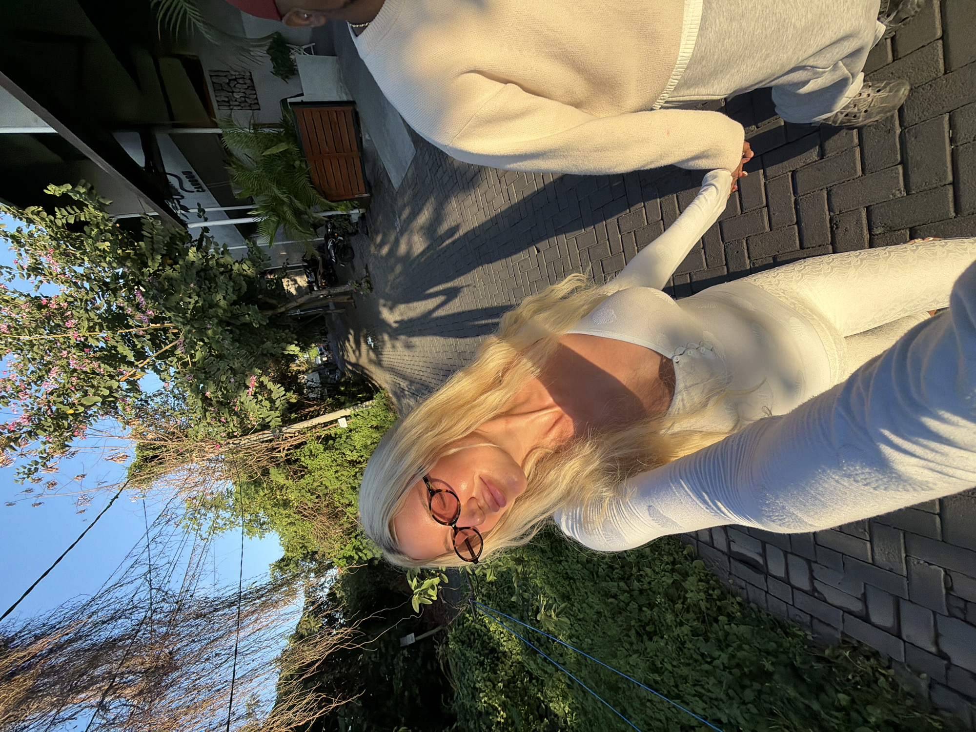 In North Kuta, Indonesia, at golden hour, a blonde woman with oval, tortoiseshell sunglasses smiles as she holds hands with a person whose body is partially visible, wearing a beige crewneck sweater and grey sweatpants. They are walking on a brick pathway lined with lush green foliage and trees. In the background, a building with a dark awning, featuring "CANOBU" signage and potted plants, suggests a commercial area. The sunlight casts long shadows across the path.