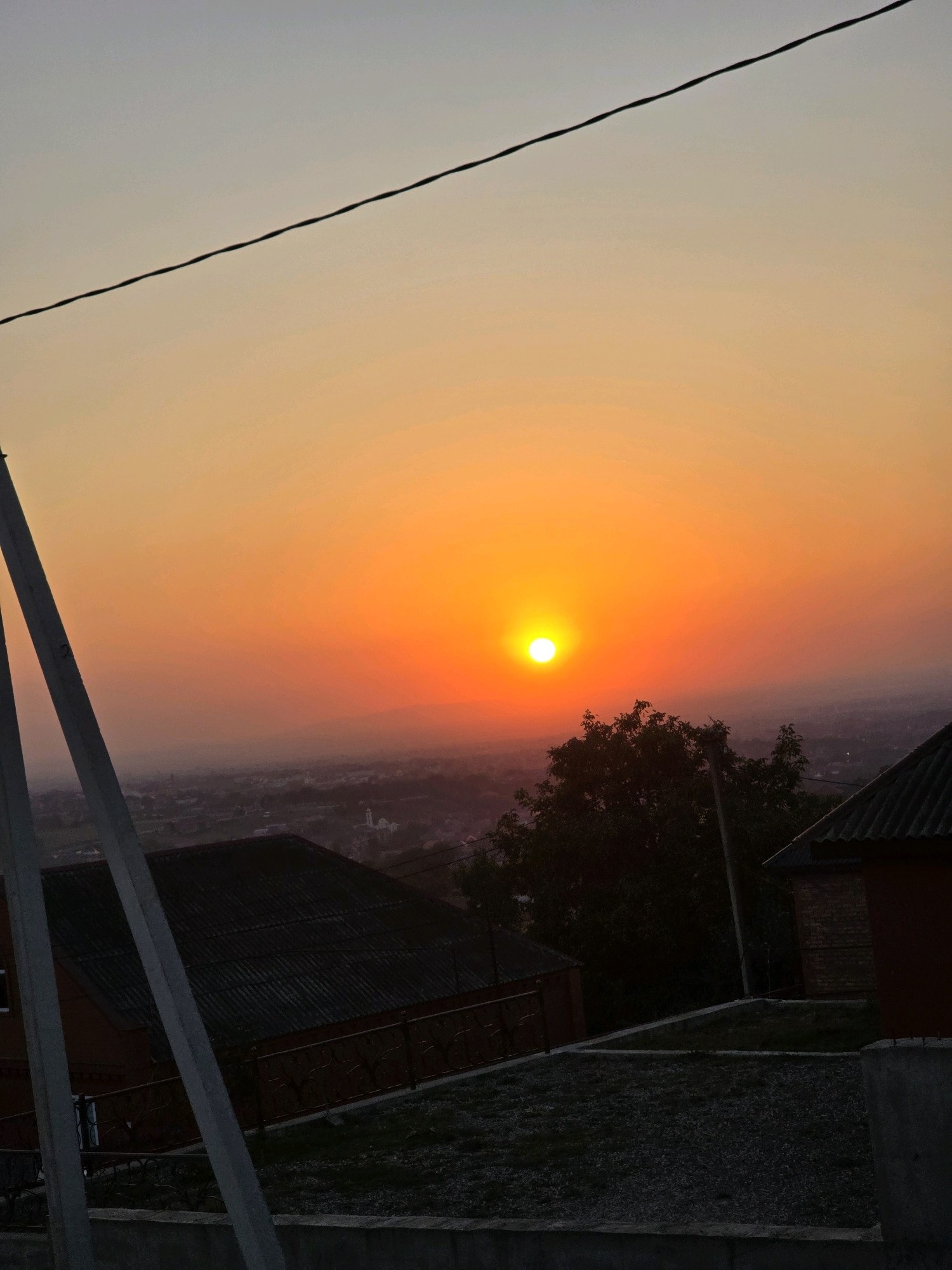 This photograph captures a sunset in the city of Alleroy, Russia. The sun, a bright orange orb, is low in the sky, casting a warm glow across a hazy landscape. In the foreground, several residential buildings with dark, corrugated roofs are visible, along with a concrete wall and some gravel. A silhouette of a tree stands to the right of the sun. In the distance, a faint outline of a minaret or tower suggests the presence of a mosque. A dark wire stretches across the top of the frame, and two utility poles are prominent on the left side of the image. The overall scene conveys a peaceful, end-of-day atmosphere.