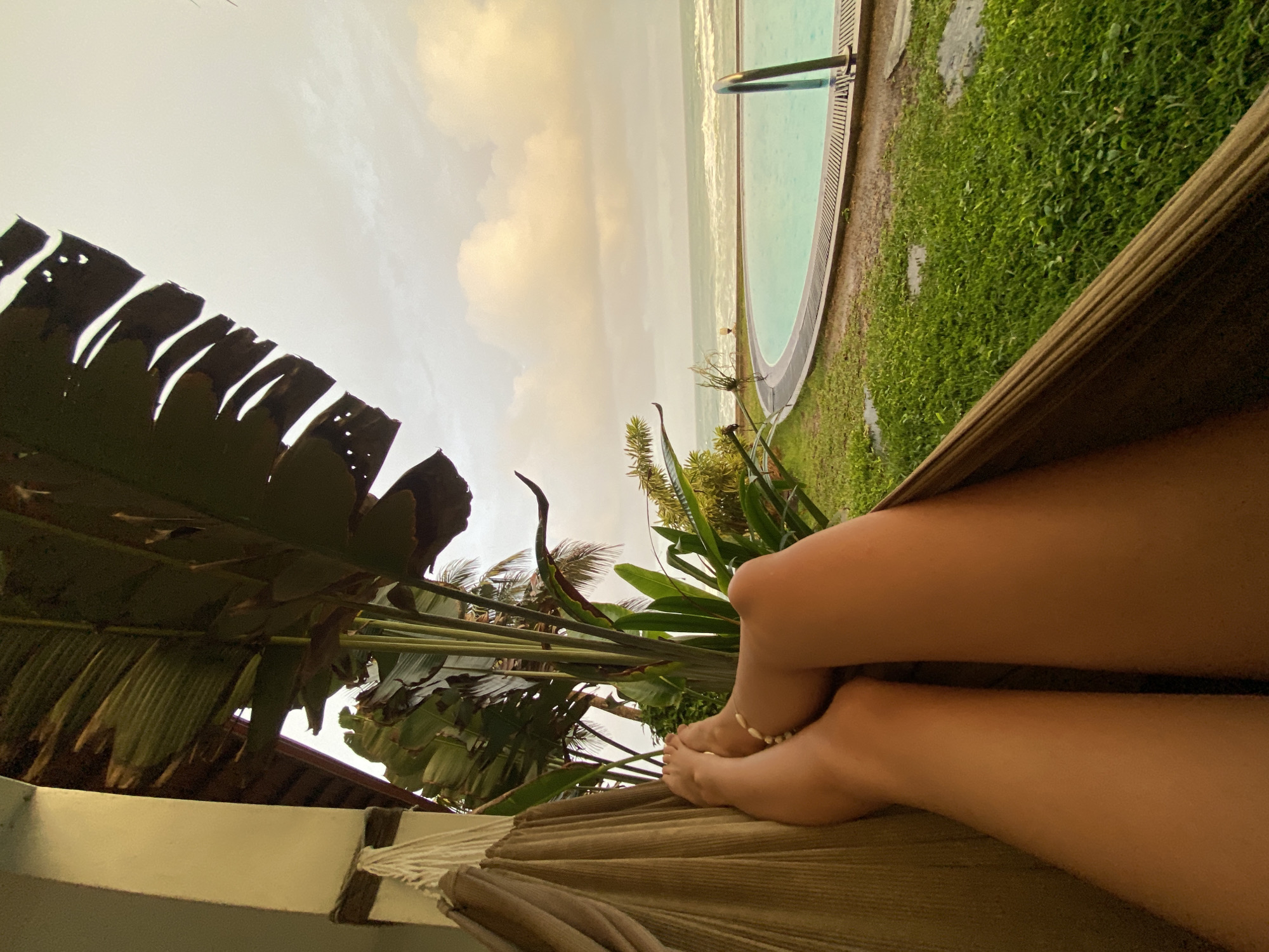 Captured from a low vantage point, the photograph presents a scene of leisure in Goviyapana, Sri Lanka. A person reclines in a hammock, legs crossed, with an anklet visible on one ankle. A dense cluster of tropical vegetation, including broad, deeply serrated leaves, frames the view. Beyond the foliage, a swimming pool edge runs parallel to a sandy beach bordering the ocean. A clouded sky completes the scene.