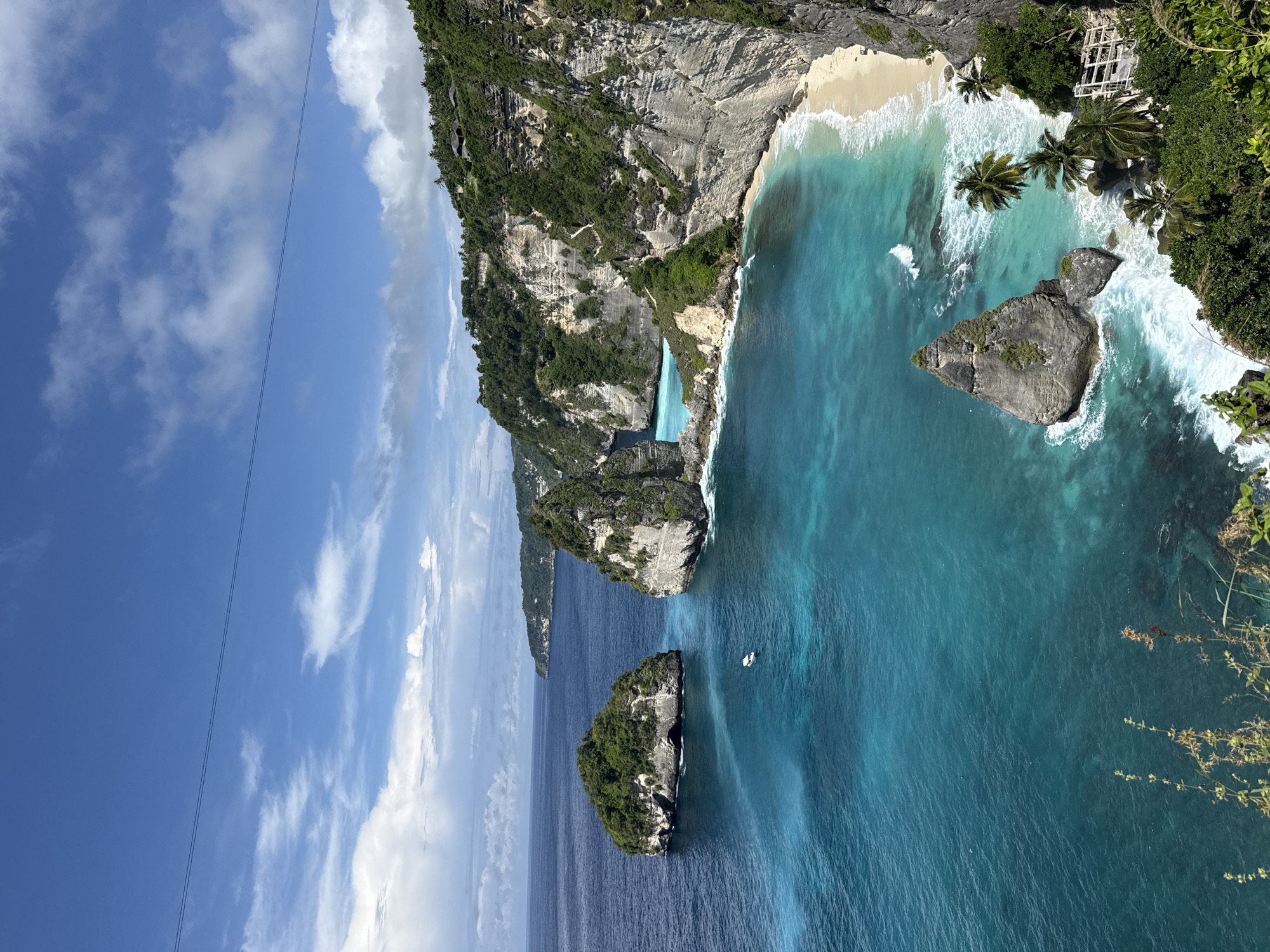 The image shows an aerial view of Kelingking Beach in Pejukutan, Nusa Penida, Indonesia. A large, jagged rock formation covered in vegetation juts out into the turquoise ocean. At the base of the formation is a white sand beach where waves are crashing. Smaller, similar rock formations are also visible in the surrounding water. Above the rock formations and ocean is a blue sky with scattered white clouds. A thin cable stretches across the sky in the upper left corner of the image.
