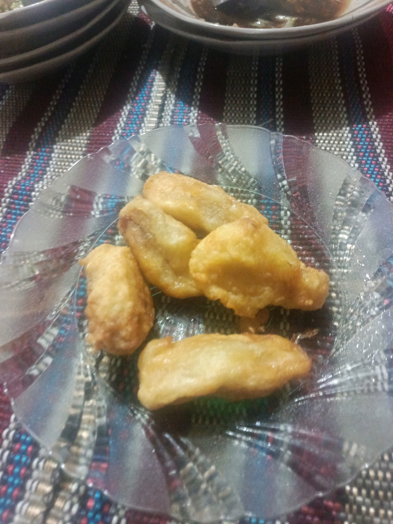 A close-up overhead shot shows a clear glass plate with a scalloped edge, filled with several pieces of fried banana. The plate sits on a woven placemat with alternating rows of dark and light threads, interspersed with thin lines of blue and red. The lighting is dim, casting shadows and making the fried bananas appear golden brown and slightly greasy. The background is out of focus, but suggests stacked plates and a dark surface.