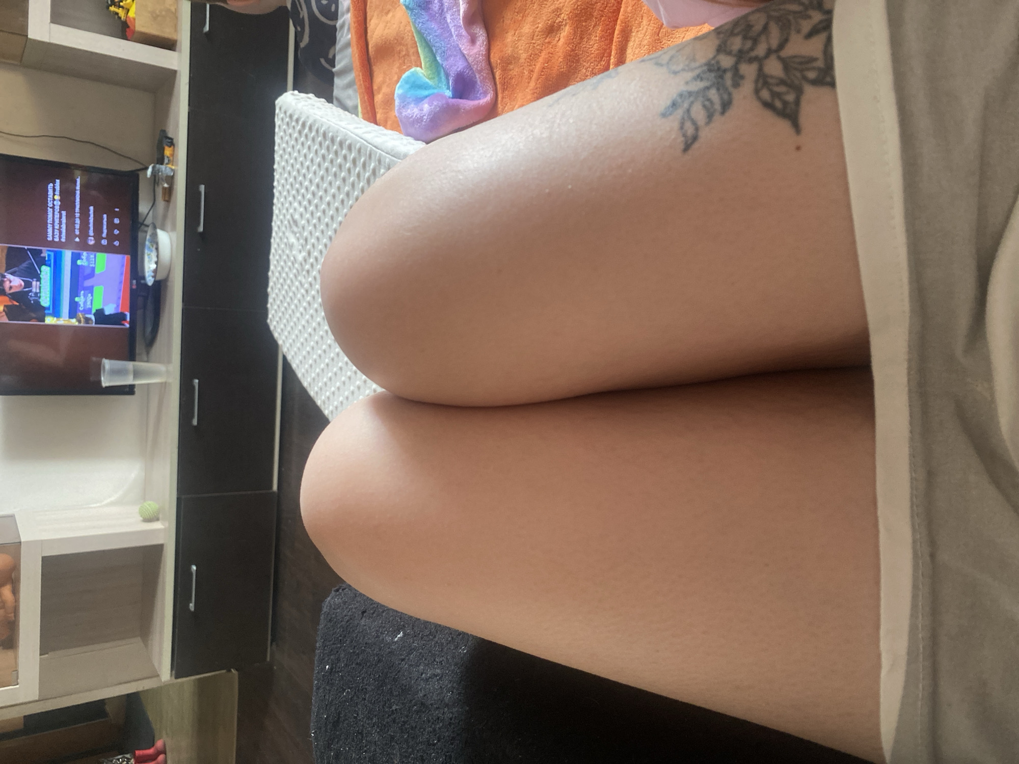 The image shows a close-up of a person's crossed legs, with a tattoo on the upper thigh of the visible leg. The person is wearing light-colored shorts or pajamas. In the background, there is a shelf with various objects and a television screen showing a video game. There is a white, textured cushion behind the legs and a dark, possibly black surface underneath the legs.
Based on the metadata, the location is Chaikovsky, Russia. However, there are no elements in the image that definitively confirm this location.