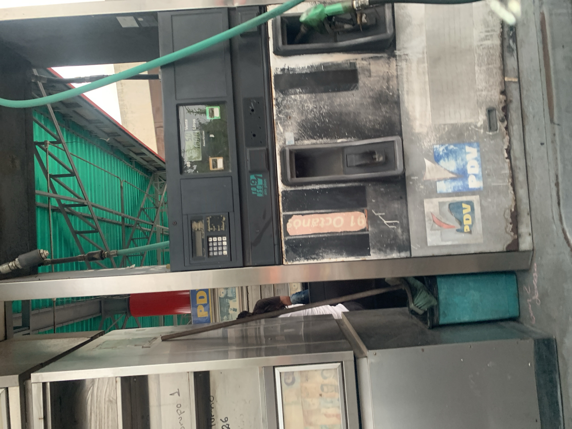 This image depicts a gas station pump and its immediate surroundings in La Fuente, Venezuela. The central subject is a heavily worn, dark gray and silver fuel dispenser, exhibiting significant dirt, grime, and rust-like discoloration. Its upper section features a digital display showing "Venta" and "Litros," with partially visible numbers suggesting "5.0" and "160.0." To the left of the main display is a numeric keypad. Below, a smaller digital readout displays "10.9" and "sin plan." An orange vertical label on the pump indicates "91 Octano." Two nozzle holsters are visible, with a green fuel hose and nozzle present in the right holster. The lower portion of the pump features two faded "PDV" logos, likely belonging to PDVSA, the Venezuelan state oil company.
To the left of the pump stands a utilitarian, metallic structure, possibly an attendant's kiosk or storage unit. Its shelves contain stacks of white items, possibly cloths or paper. Handwritten labels such as "grupo 1," "turno," and "26" are affixed to its surface. A framed display containing what appears to be currency or small pictures is visible on the lower section of this structure. Partially obscured between this structure and the pump, an individual wearing a white t-shirt and dark pants is visible from the back, facing away from the camera.
At the base of the pump and the adjacent structure, a turquoise cylindrical container, likely a trash bin, is situated. A broom with a green head and a long handle rests beside it. The overhead environment features a green corrugated metal canopy supported by a dark metal truss structure. A bright red cylindrical object is visible beneath the canopy, partially behind the pump. The ground beneath is concrete or asphalt, with faint, illegible writing present. The overall scene conveys a sense of operational use despite visible wear and lack of pristine maintenance.