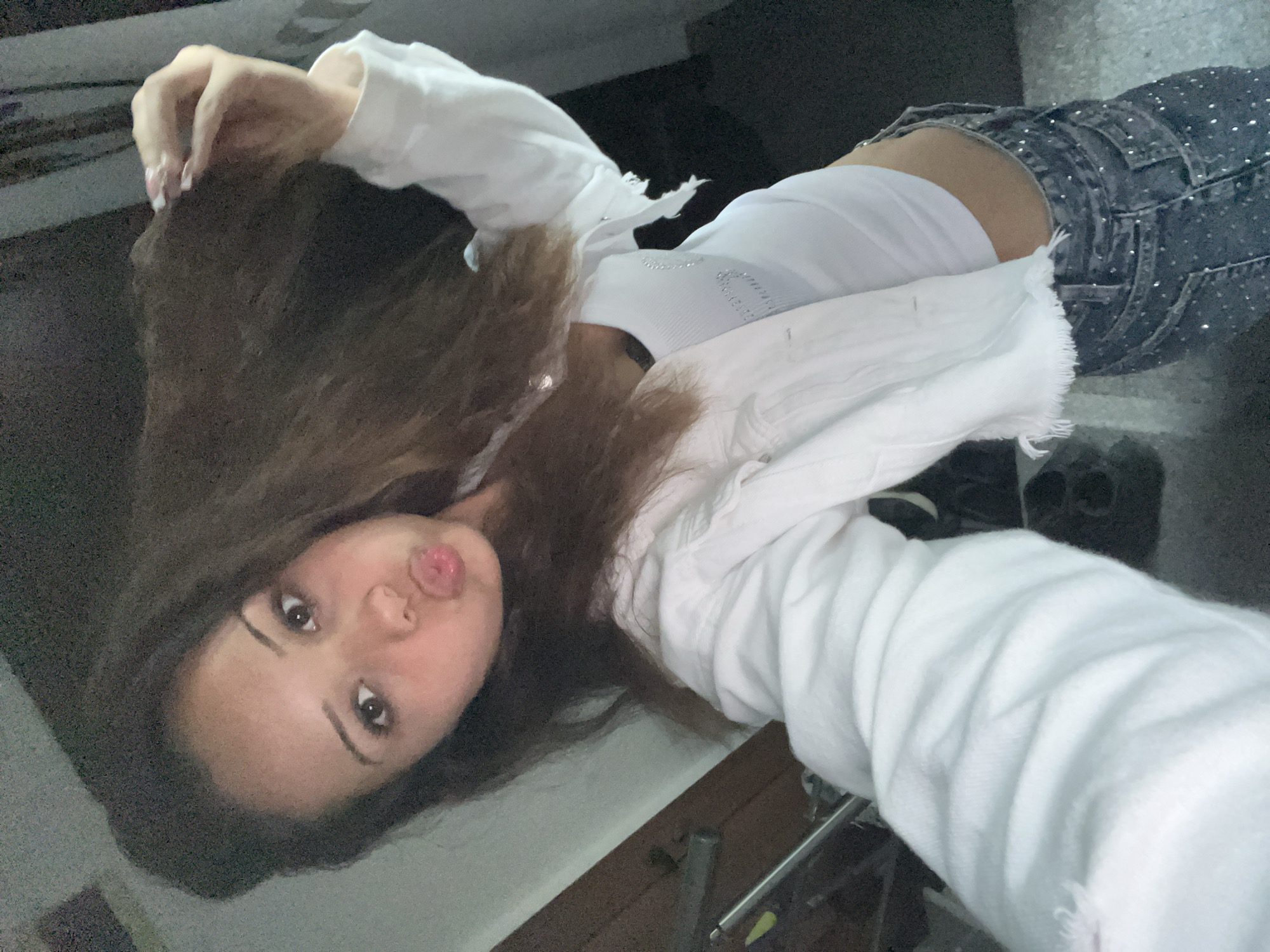 A single female subject is depicted in an indoor selfie, captured from a slightly elevated perspective. The subject, appearing to be in her late teens or early twenties, has long, light brown hair styled loosely. She is making a "duck face" expression with puckered lips and maintains direct eye contact with the camera. Her right hand is raised to the upper right side of her head, with fingers splayed, appearing to interact with her hair; her fingernails are manicured with white tips. She wears a white, long-sleeved denim jacket with a frayed hem, layered over a white ribbed crop top that features subtle, possibly rhinestoned, embellishments on the chest. Her lower garment consists of dark-wash jeans extensively adorned with small, reflective silver studs or rhinestones. The background is dimly lit and indistinct, showing dark, possibly wall or furniture elements to the left and rear, and a dark floor. The setting is located in the city of Unknown, Unknown.