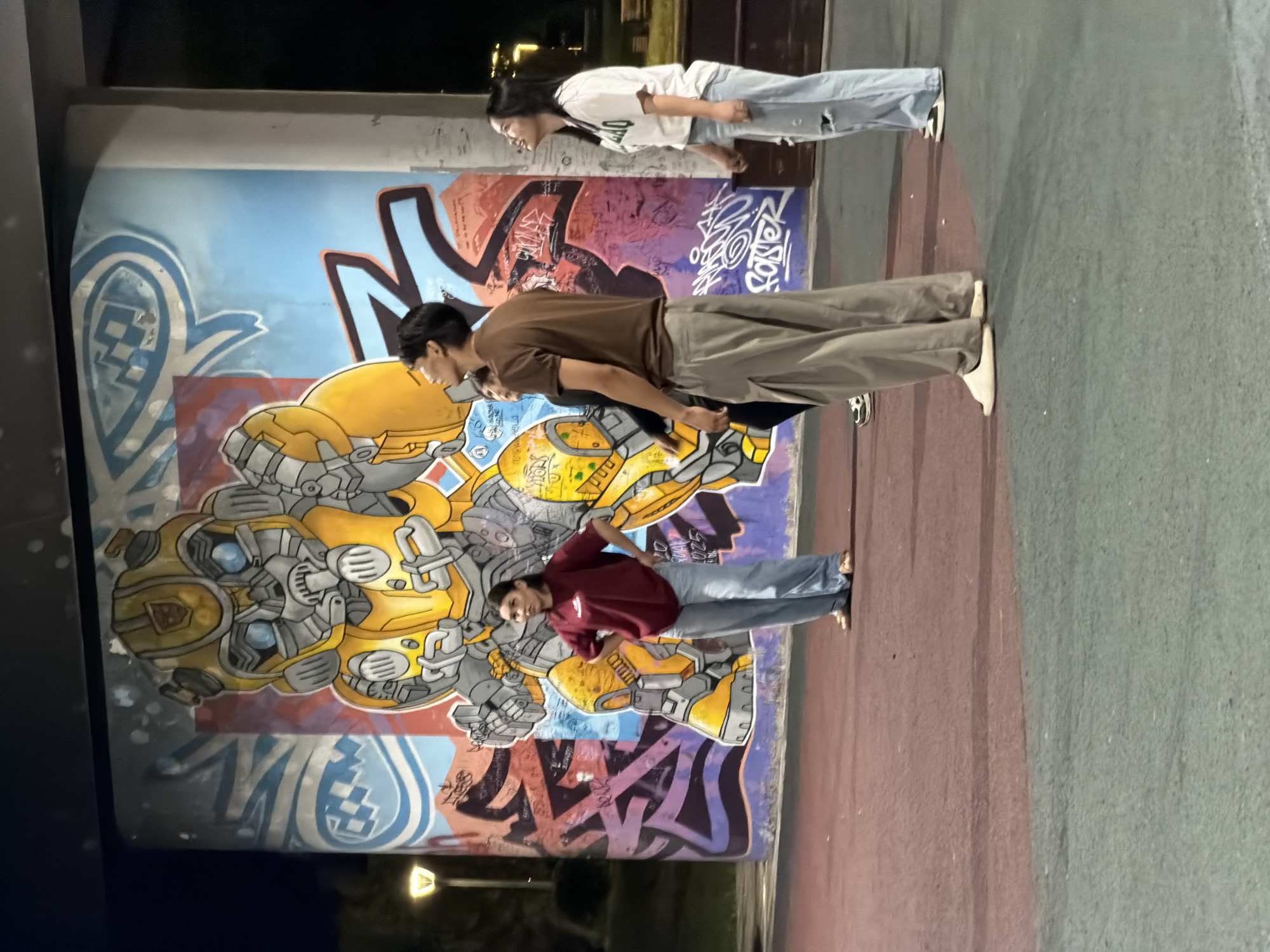 This is a medium shot, taken outdoors at night, of a group of four young people standing in front of a large, colorful mural. The mural depicts Bumblebee from the Transformers franchise, with bright yellow and grey colors dominating the character's design. The background of the mural is a vibrant blue, with graffiti in various colors. The ground in front of the mural is a reddish-brown, with a green section visible in the lower part of the frame.
The four individuals are standing at varying distances from the mural and each other, appearing to be looking up at it. From left to right:
1. A young woman wearing a maroon t-shirt and light blue jeans. She is standing with her hands on her hips, looking slightly to her left.
2. A young man wearing a brown t-shirt and loose-fitting grey pants. He is looking upwards towards the mural, with his head tilted back.
3. A young woman wearing a white t-shirt with green writing and light blue ripped jeans. She is looking upwards and to her right, with her head tilted back.
4. A young woman wearing a white t-shirt and light blue jeans. She is looking upwards towards the mural.
A street lamp is visible in the background to the left of the mural. The overall scene suggests a casual gathering of friends admiring street art in an urban environment. This scene is located in Astana, Kazakhstan.