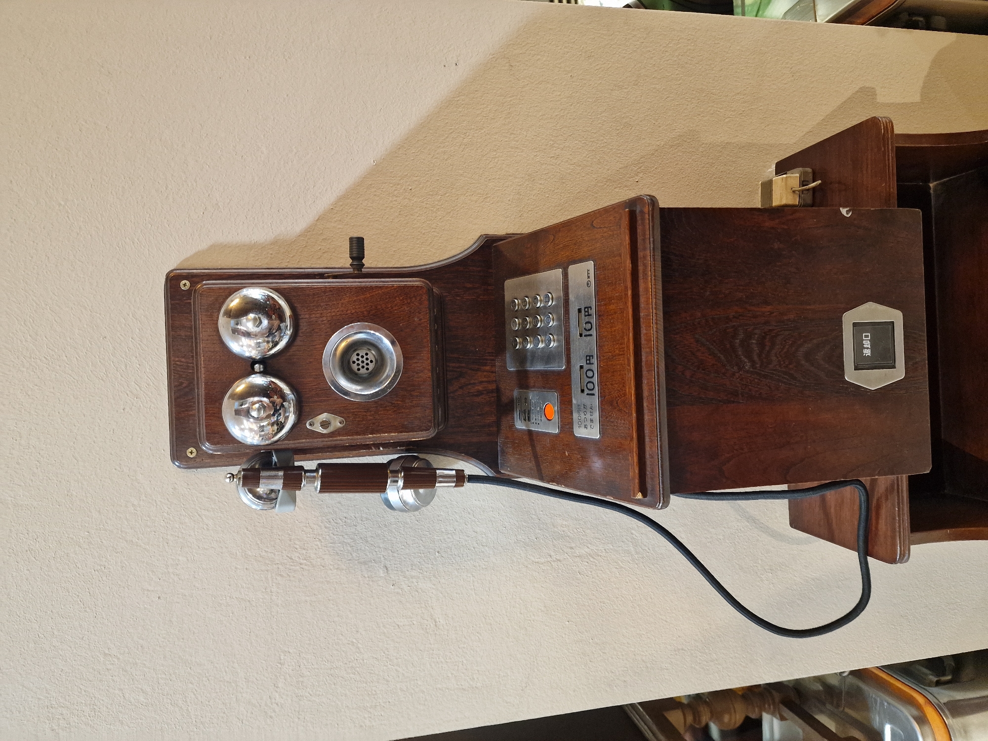 This media file showcases a vintage wooden payphone mounted on a textured, light-colored wall. The payphone is the central object, featuring a dark wood casing with polished metal accents. On the left side of the main body, there are two prominent, circular, silver bells. Below the bells, a wooden and silver-toned handset is attached via a black coiled cord.
To the right of the bells, a circular metal grille is visible, likely an intercom or speaker. Above this grille, a small, dark knob protrudes from the wood. Further to the right, a panel with a keypad and a slot for coins is integrated into the phone's design. The keypad has a metallic finish, and a small, orange button is visible below it. To the right of the keypad, there's a metallic slot with Japanese text, indicating it's a coin slot.
The visible text on the payphone reads: "100円は おつりが でません 100円," which translates to "100 yen, no change will be given." Another line reads "返却口" (henkyakuguchi), meaning "return slot," and "10円" (10 yen). A small black switch is also visible on the far right of the phone's body, labeled with Japanese characters.
The lighting suggests an indoor setting, possibly a public space or a museum, given the preserved nature of the vintage item. The shadow cast on the wall indicates a light source from the top left. No people are present in the frame, and the scene focuses solely on the antique telephone.