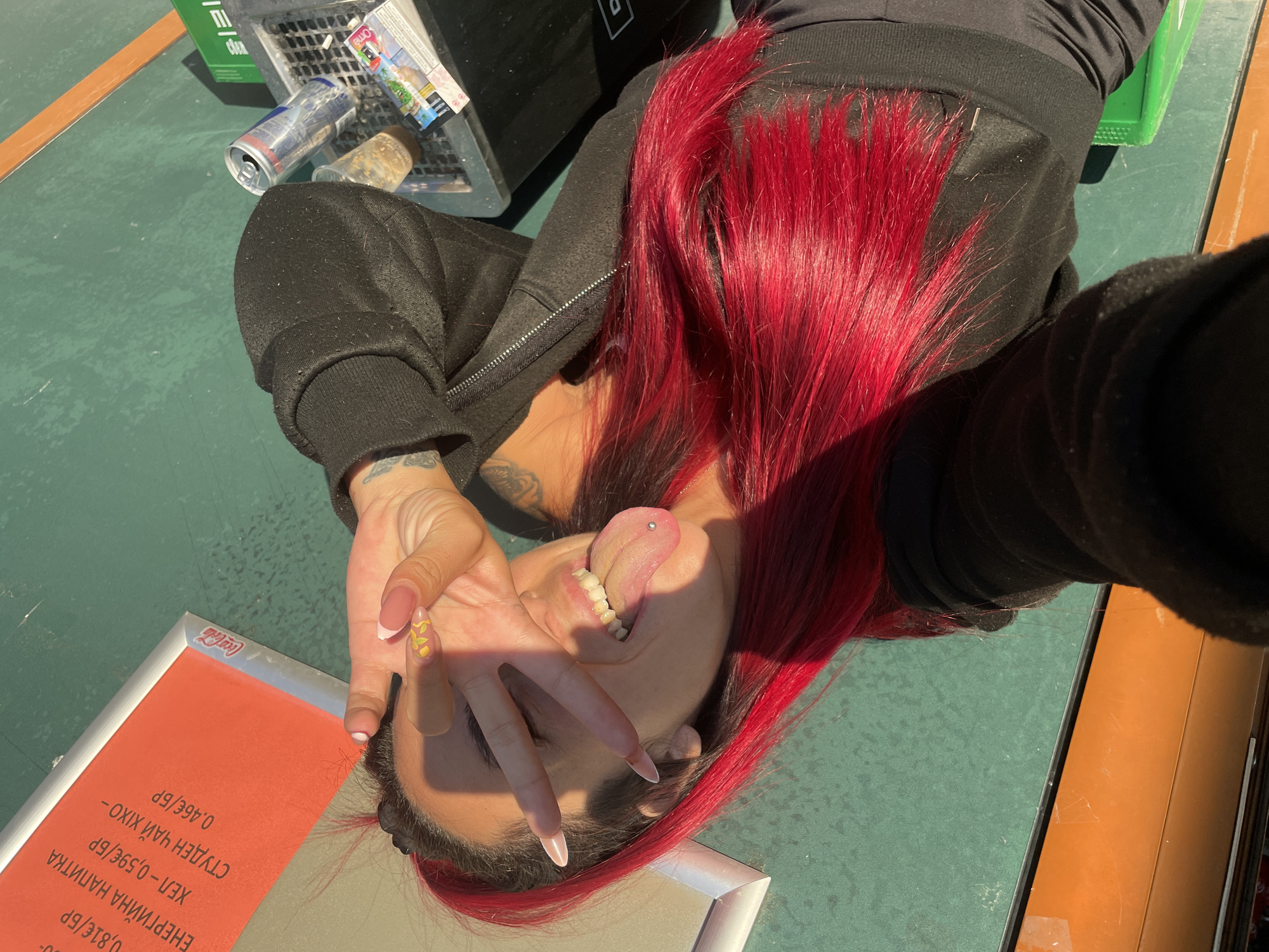 A young person with bright red hair and a tongue piercing is lying on their side, looking at the camera. Their right hand is raised, with long, manicured nails painted with a French tip and a small yellow design on one nail. They are wearing a black zip-up hoodie. The background shows a green surface, likely an outdoor table or court, with a trash bin in the upper left corner containing cans and packaging. An orange surface runs along the right side of the frame. A sign with text in Bulgarian, including prices in Euros, is visible in the lower left corner, along with a Coca-Cola logo. The lighting suggests it is daytime.