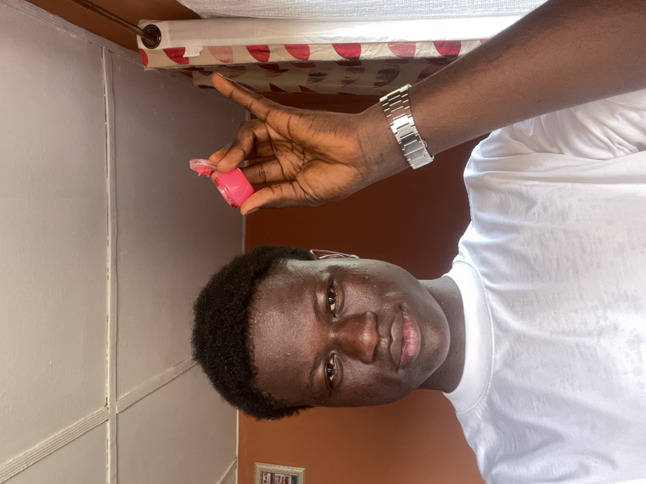 An indoor, waist-up portrait captures a young adult male with dark skin and short, dark, curly hair, facing directly forward. He wears a plain white crew-neck t-shirt and a silver-toned metal watch on his left wrist. His right hand is raised, holding an open, small, circular, bright pink container, likely a lip balm, above his head to the right. The background features an orange-brown wall to the left, with a partially visible framed picture. To the right, a window is covered by a sheer white curtain, behind which a patterned curtain with red and white circular motifs hangs from a brown rod. The ceiling is white with visible structural lines. The scene is located in Mosafejaw, Nigeria.