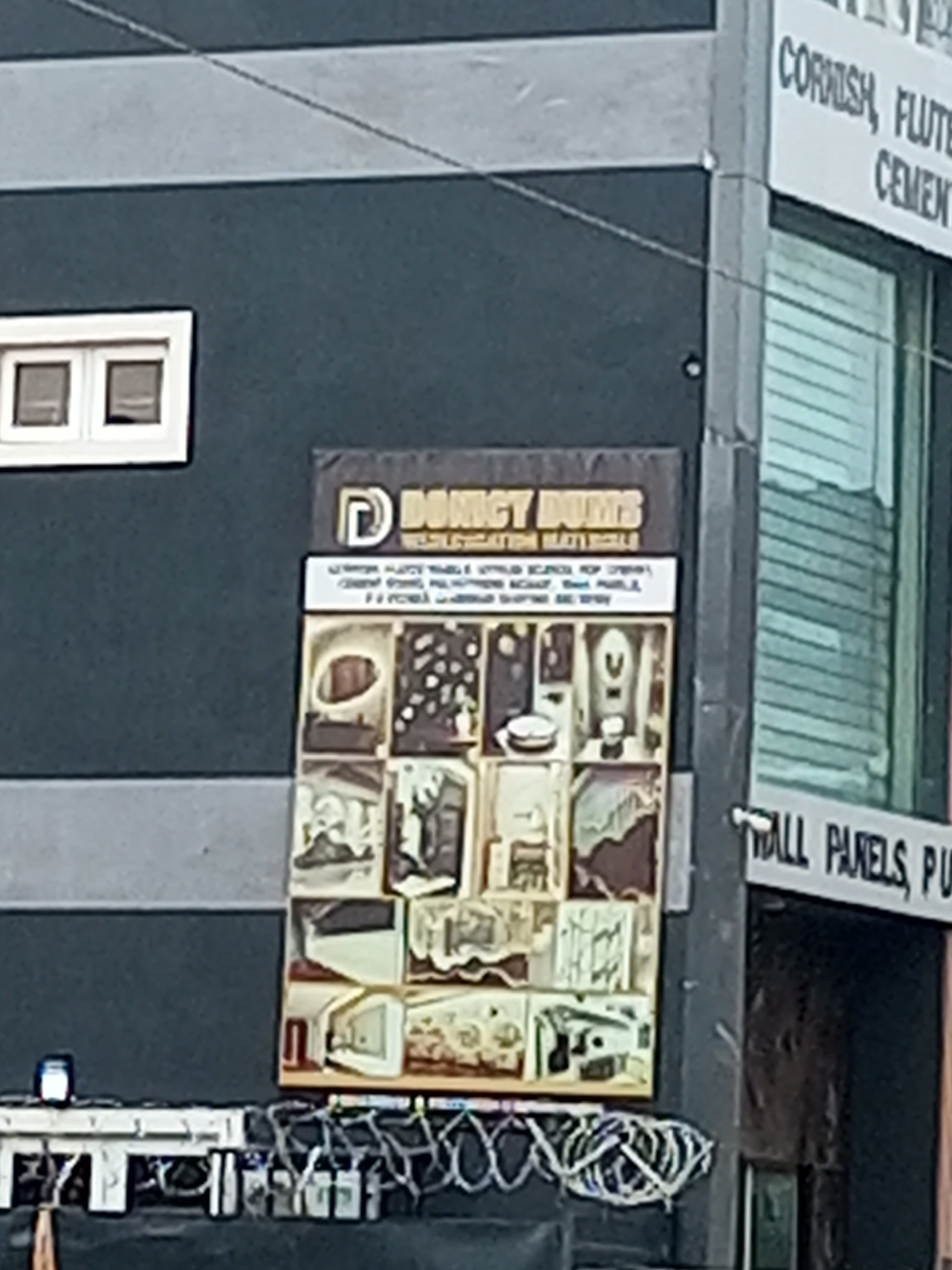 A partial view of the exterior of a multi-story building in Maiburiji, Nigeria, is shown. The building's facade is painted in gray and a darker shade of blue. A sign is mounted on the side of the building, bearing the name "DOMICY DUMS" and the phrase "INTERIOR DECORATION MATERIALS". The sign also has a grid of images that appear to be examples of the company's work or products. Below the sign, there is a ledge containing what appears to be various cables and wires. The side of the building has a window with white trim and horizontal blinds. Adjacent to the structure with the sign, another part of the building is visible, featuring a sign that partially reads "CORNISH, FLUTE... CEMENT..." and "WALL PANELS, PU...". The scene is captured in daylight with a somewhat grainy quality.