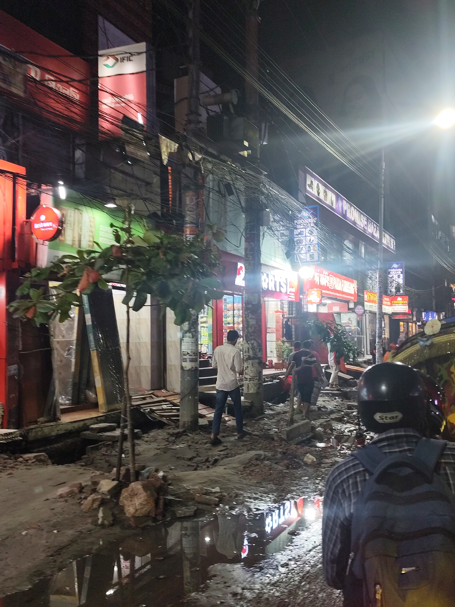 Night scene in Dhaka, Bangladesh. A street is partially destroyed with rubble and standing water. Pedestrians walk on the damaged street. Overhanging electrical wires are visible above shops and storefronts with bright signs, including "IFIC" and "PROMISE BAZA". A person wearing a helmet and backpack is in the foreground.