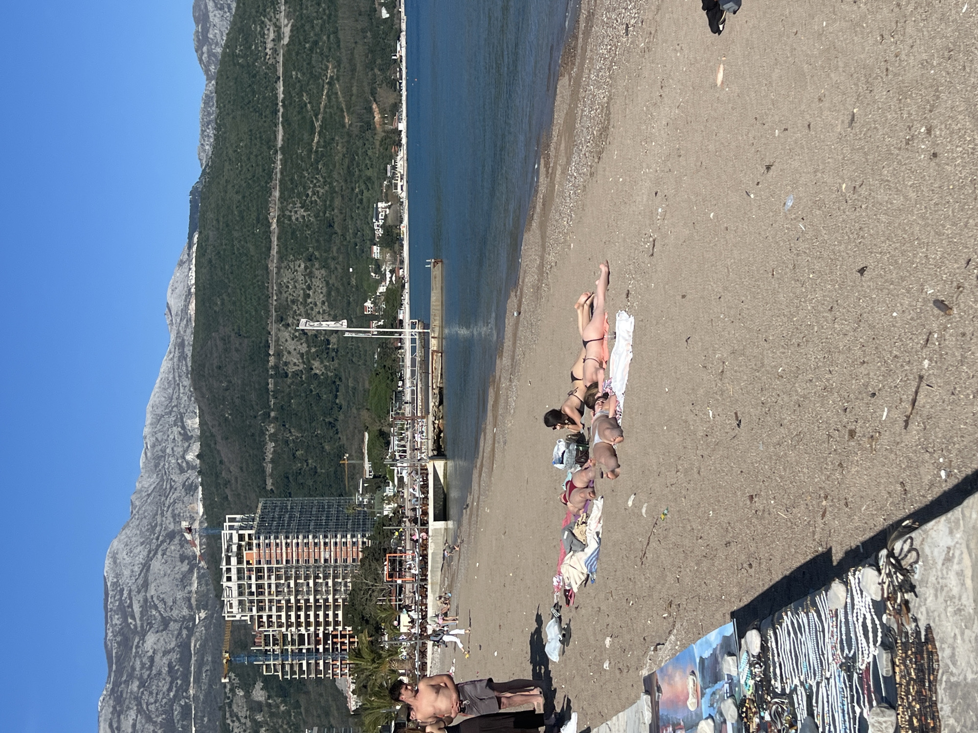 The image shows a beach scene in Budva, Montenegro. A mountainous landscape with green vegetation and some buildings rises sharply on the left side of the image. A tall, unfinished building stands closer to the beach. The blue sea extends horizontally across the upper part of the image.
On the sandy beach, several people are sunbathing and relaxing. Some are lying on towels, and a few are gathered near the shoreline. The foreground features a display of jewelry and artwork set up near a concrete ledge, implying a vendor or a market. A man stands shirtless, with other people nearby. The overall impression is a sunny day at a tourist beach.