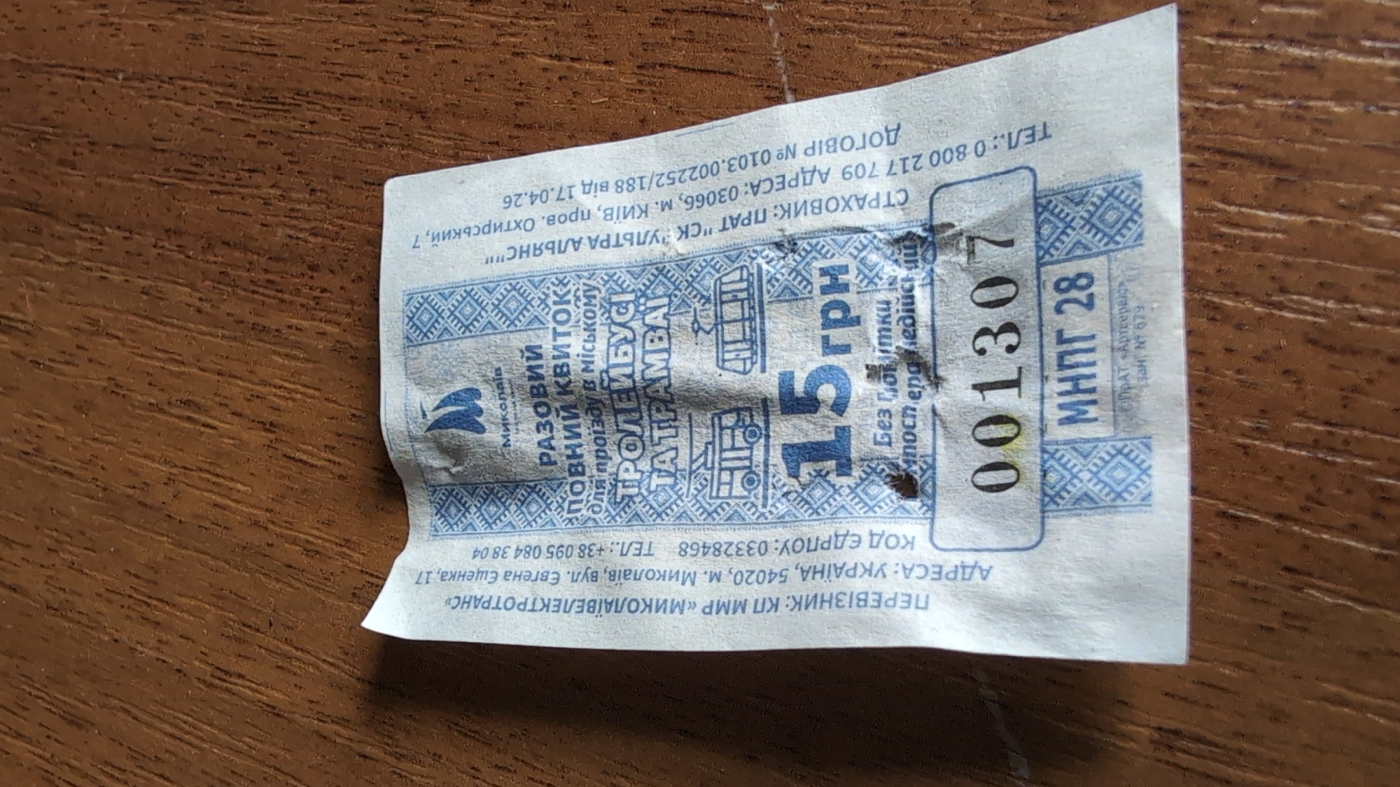 The image shows a close-up, top-down view of a crumpled paper ticket on a wooden surface. The ticket is light blue and white with intricate patterns. Text on the ticket is in Ukrainian. The ticket appears to be for public transportation in Mykolaiv, Ukraine.
Key details on the ticket include:
* "ПЕРЕВІЗНИК: КП МMP «МИКОЛАЇВЕЛЕКТРОТРАНС»" which translates to "CARRIER: KP MMP "MYKOLAEVELECTROTRANS"". This indicates the transport company.
* "АДРЕСА: УКРАЇНА, 54020, м. Миколаїв, вул. Євгена Єщенка, 17" which translates to "ADDRESS: UKRAINE, 54020, Mykolaiv, st. Yevhena Yushchenka, 17". This is the specific address of the transport company in Mykolaiv.
* "ТЕЛ.: +38 095 084 38 04" which is a phone number.
* "РАЗОВИЙ ПОВНИЙ КВІТОК" which translates to "SINGLE FULL TICKET".
* "ДЛЯ ПРОЇЗДУ В МІСЬКОМУ ТРОЛЕЙБУСІ ТА ТРАМВАЇ" which translates to "FOR TRAVEL IN CITY TROLLEYBUS AND TRAM".
* A price of "15 грн" which means 15 Ukrainian Hryvnia.
* A ticket number "001307".
* Below the ticket number is "МНПГ 28".
* There are also details of the insurance company: "СТРАХОВИК: ПРАТ "СК "УЛЬТРА АЛЬЯНС"" and an address "м. КИЇВ". The contract number and date are also visible.
The ticket is slightly creased and has a small yellow discoloration in the number "001307". The wooden surface is a dark brown with a visible grain pattern.