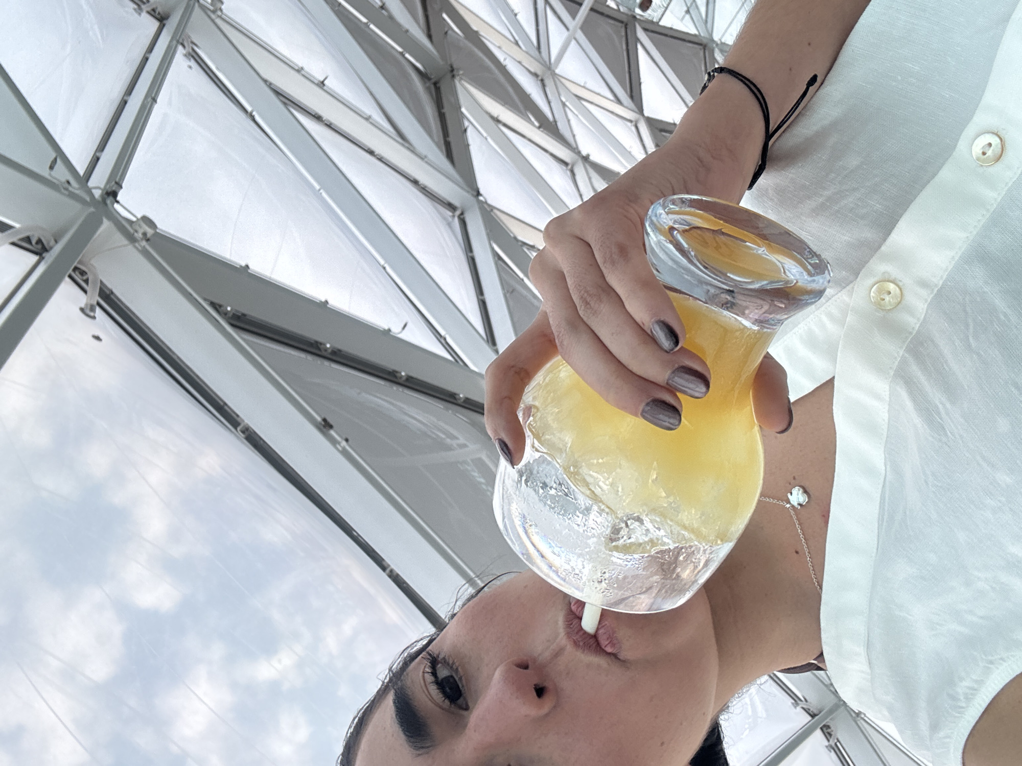 A woman drinks a yellow drink in a glass with a straw. She is wearing a white shirt, and there is a diamond necklace around her neck. The woman is holding the glass with her right hand, and she has gray nail polish. The background is a geometric building structure.