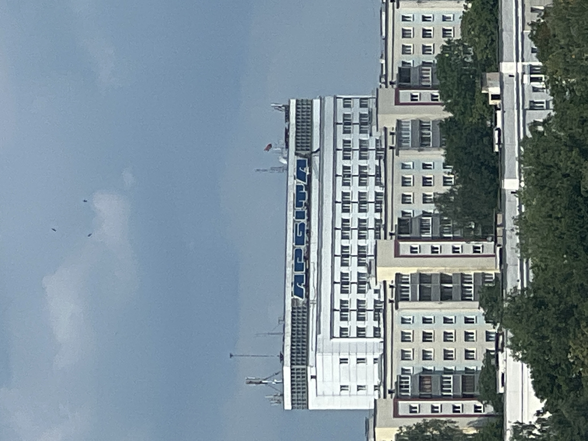 The image shows a view of the Belarusian State University of Informatics and Radioelectronics (BSUIR) in Minsk, Belarus. The building is tall and white, with a prominent sign that reads "APCSITA" in blue letters. The building has many windows, and there are several antennas on the roof. In front of the BSUIR building, there are several residential buildings, which appear to be apartment complexes. These buildings are multicolored, with sections of yellow, blue, and gray. There are trees in the foreground, obscuring the lower portions of the residential buildings. The sky is a pale blue with some clouds. There are also three small dark objects in the sky, which could be birds or insects.