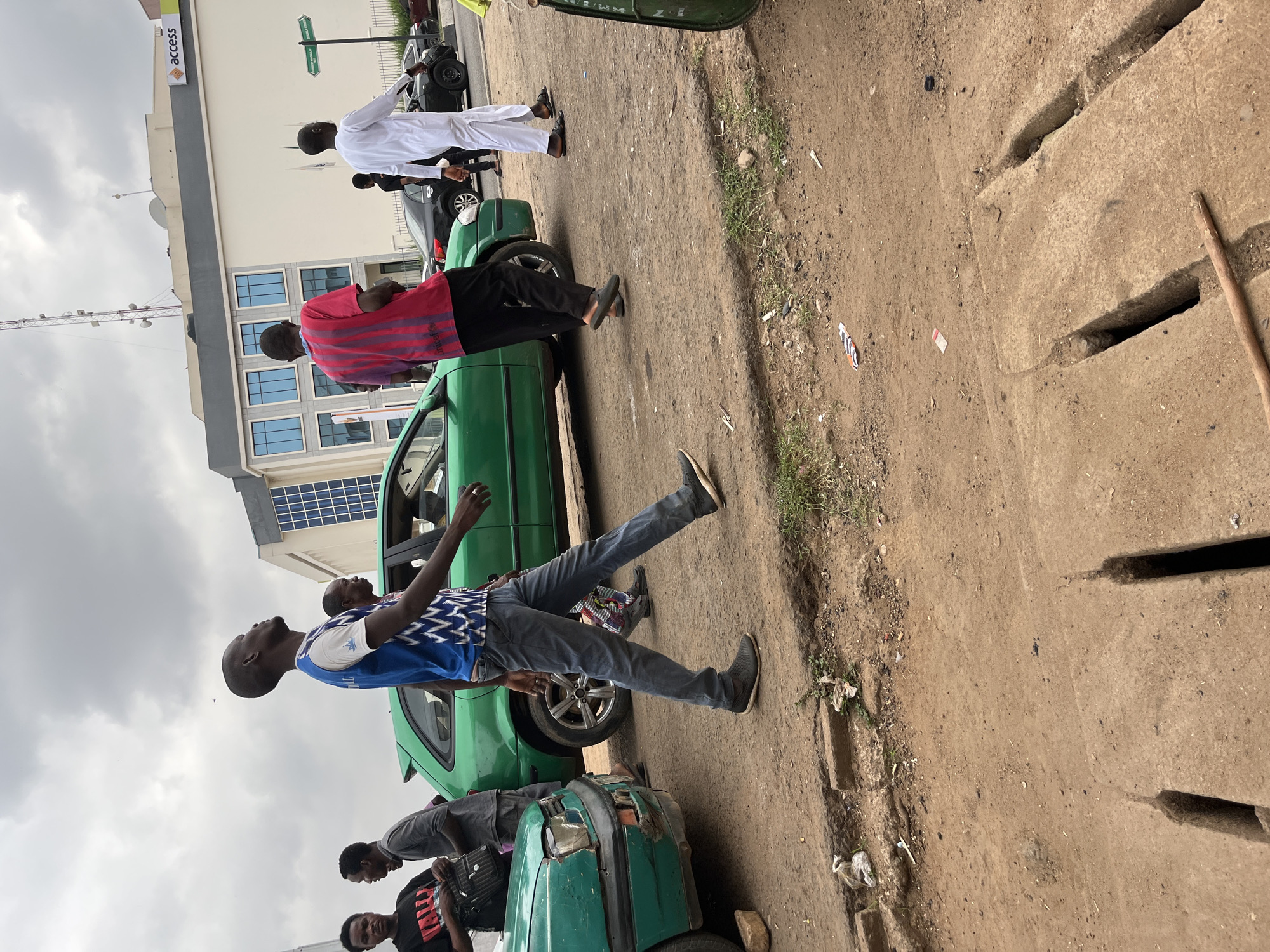 The image depicts a street scene in Abuja, Nigeria. Several people are standing near green cars. Some are walking along the sidewalk, while others are positioned near or leaning against the cars. The environment includes a building with "access" signage, indicating a potential bank location. The road shows signs of wear, with cracks and debris visible. There are concrete drainage channels along the edge of the road. The sky is overcast, suggesting a cloudy day.