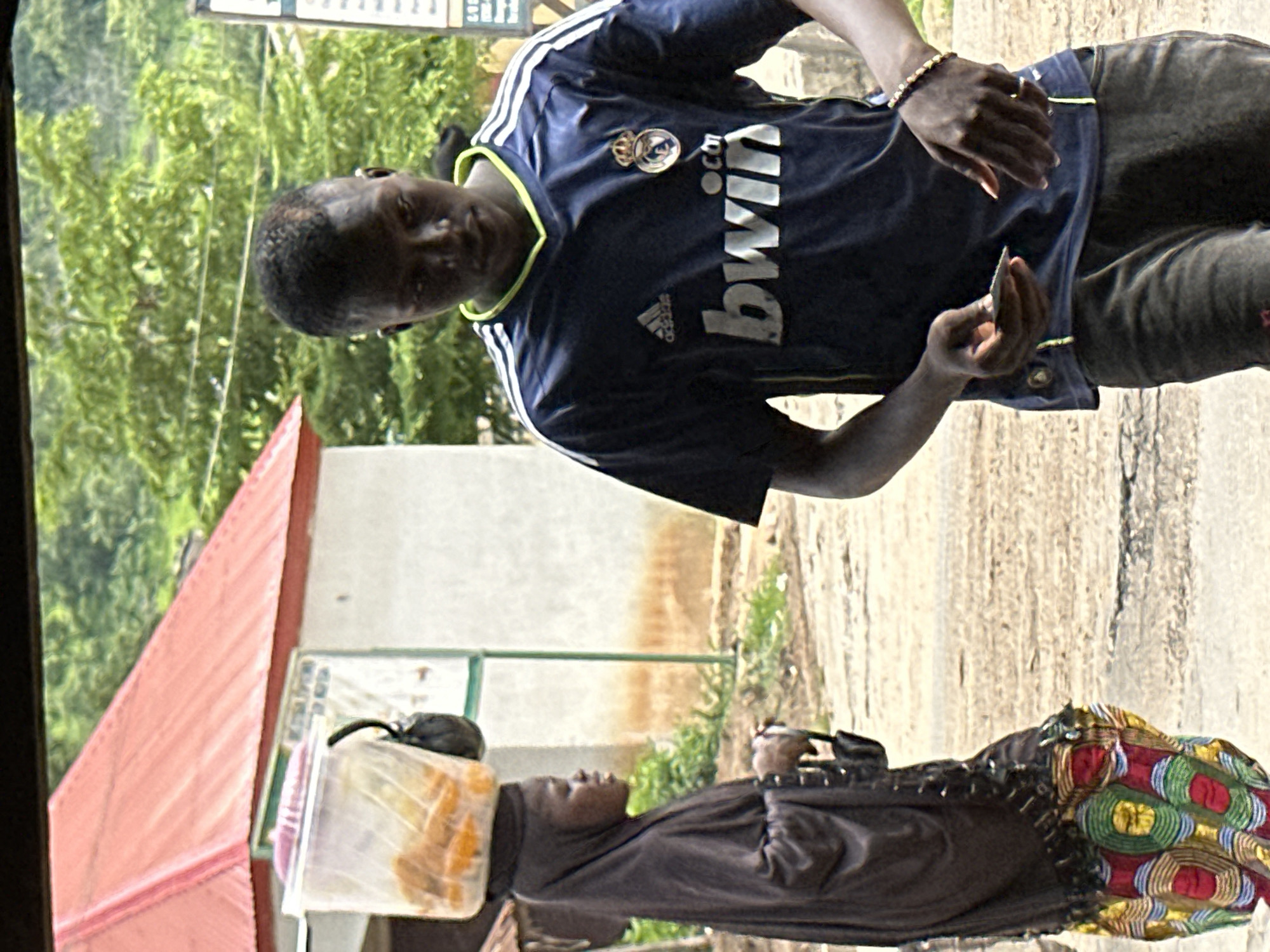 A Nigerian man wearing a blue Real Madrid jersey with "bwin" printed across the chest and a woman in a dark hijab and a colorful skirt are seen on a dirt road in Mararaba, Nigeria. The man holds a small object, possibly a phone or card, in his left hand and wears a beaded bracelet on his right wrist. The woman has a clear plastic container filled with what appears to be food items balanced on her head, and a dark bag is attached to the side of the container. Behind them, a building with a red corrugated metal roof and white walls is visible, surrounded by green foliage. The scene depicts an outdoor marketplace or roadside setting.